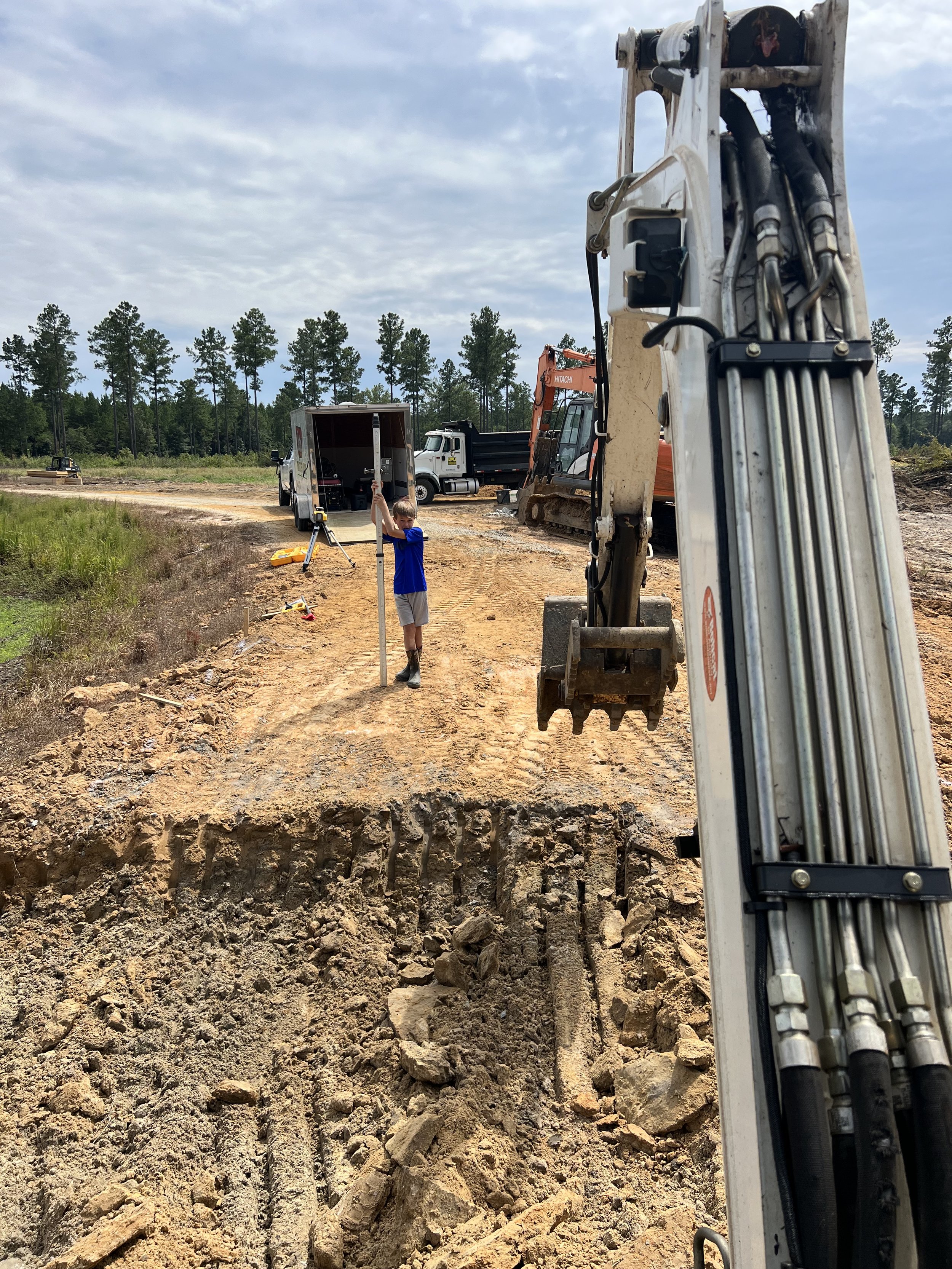 A construction site with a boy holding a measuring stick, construction machinery, and trucks on a dirt road, with a forest in the background.