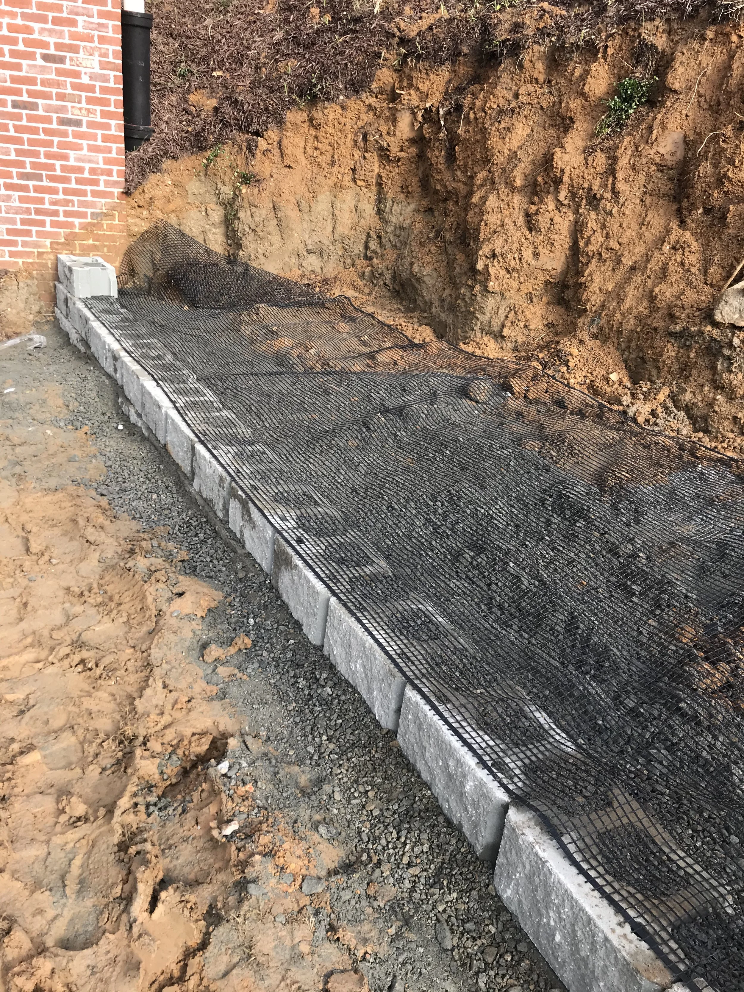 Construction site with a new concrete sidewalk bordered by concrete blocks, black mesh reinforcement, and a brick building on the left side.