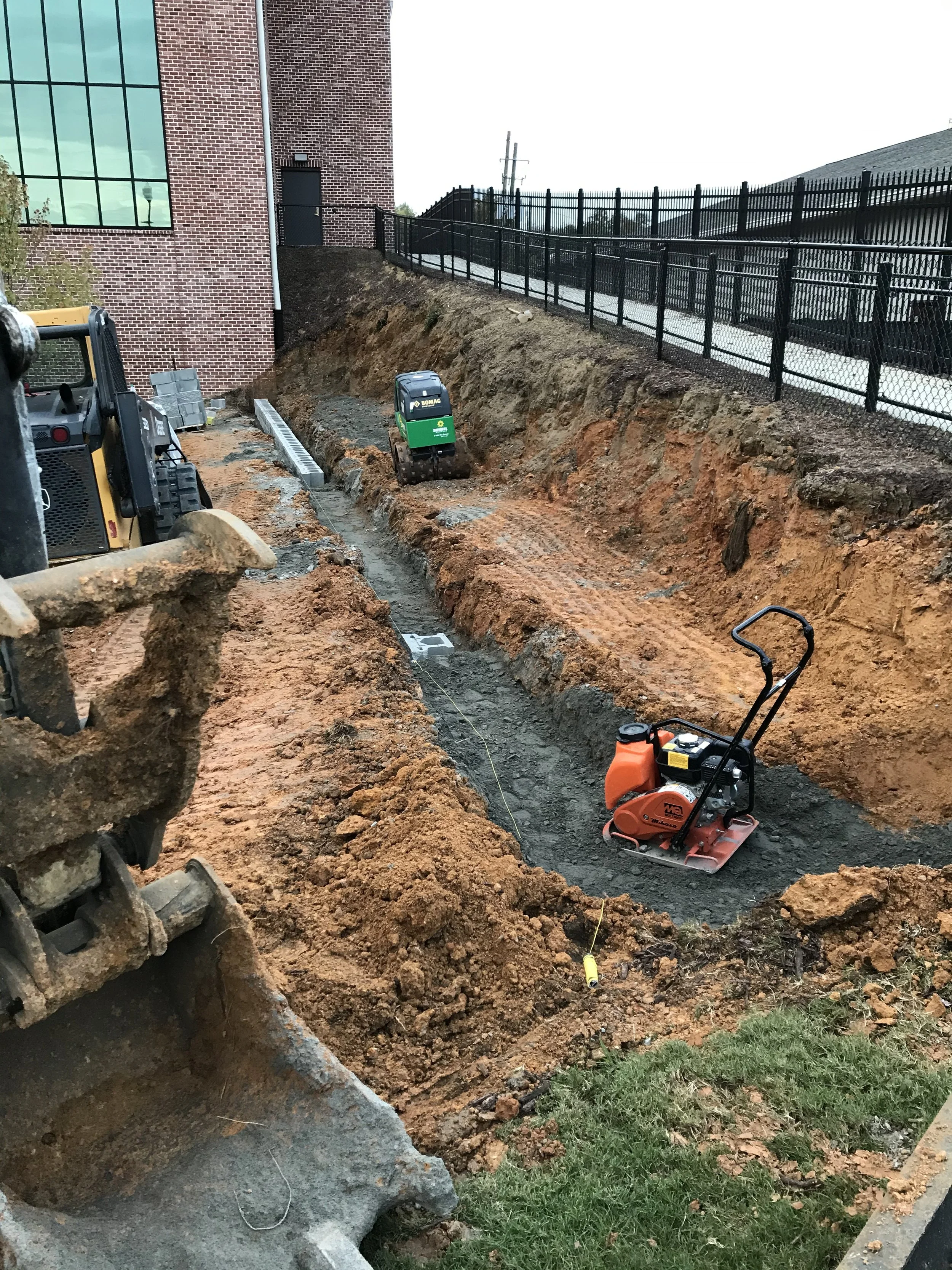 A construction site showing a deep trench with equipment including a small compactor and a mini excavator. The site is next to a brick building and fenced area.