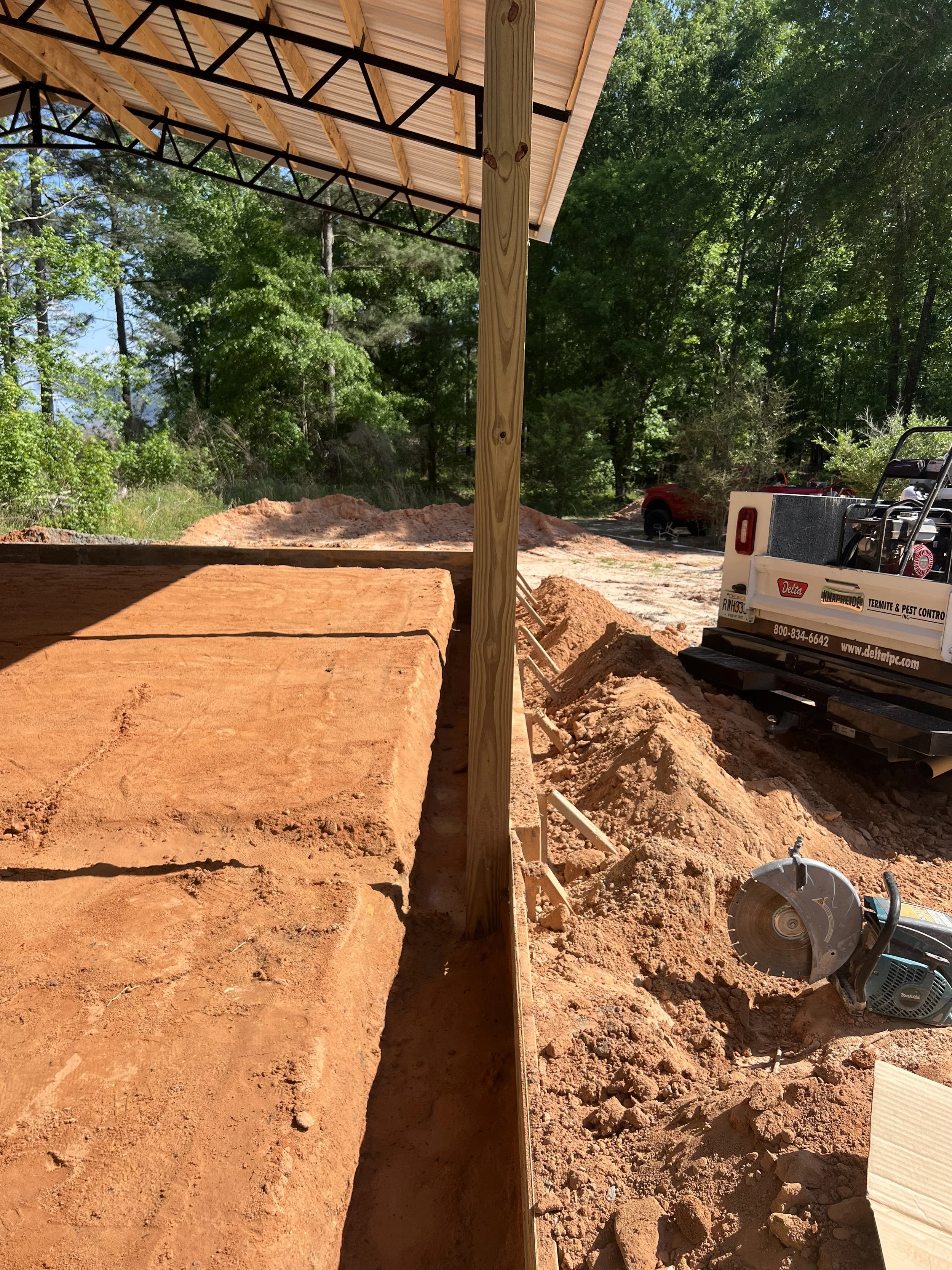 Construction site with dug trenches, a wooden post supporting a roof, and construction tools including a circular saw. There are trees and a vehicle in the background.