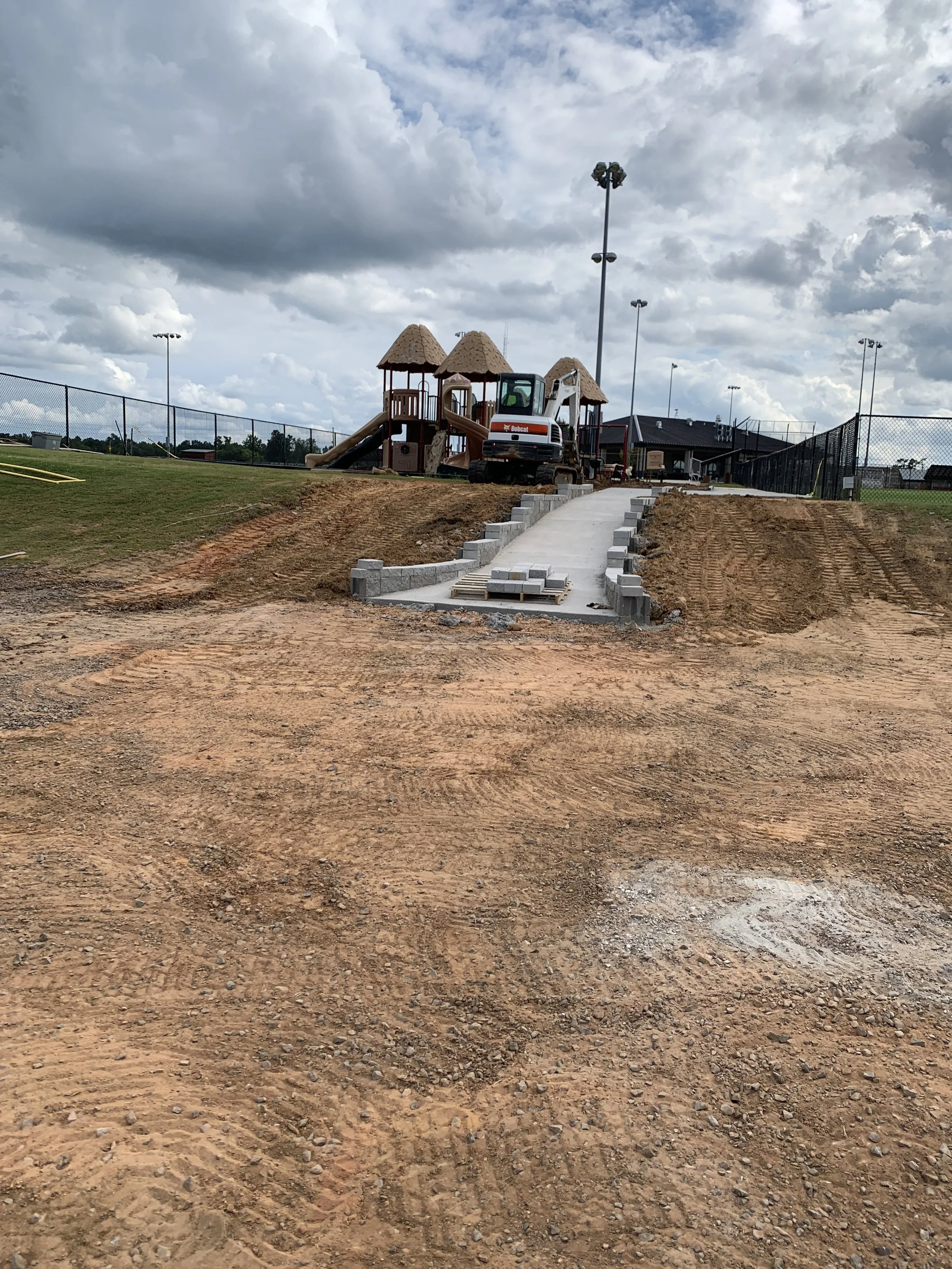 A playground under construction with a concrete pathway, a small digger, and a play structure with thatched roof huts, surrounded by a fence and athletic fields, under cloudy skies.
