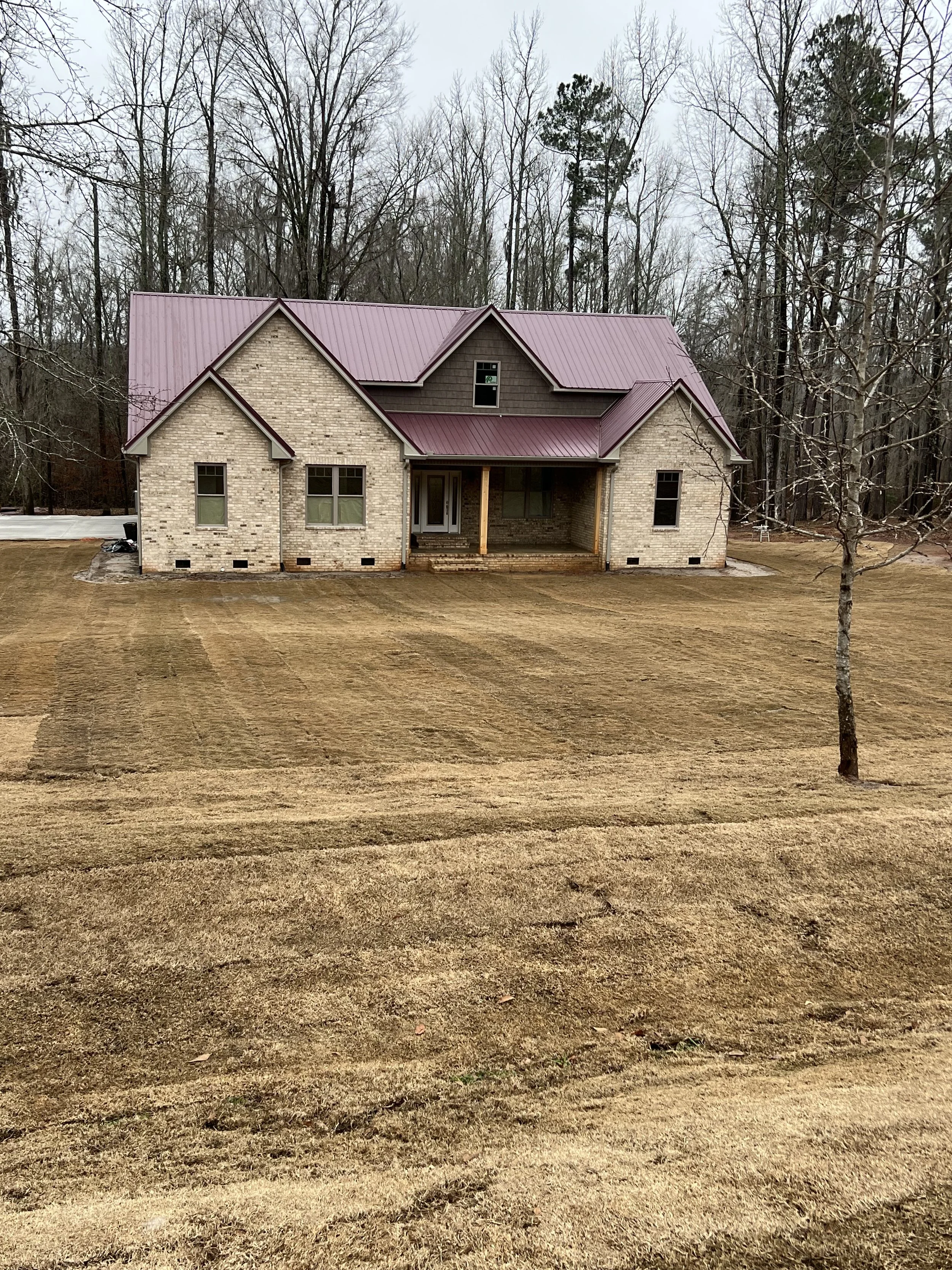 Newly constructed two-story brick house with a maroon metal roof, front porch with stairs, surrounded by a grassy lawn and trees in the background.