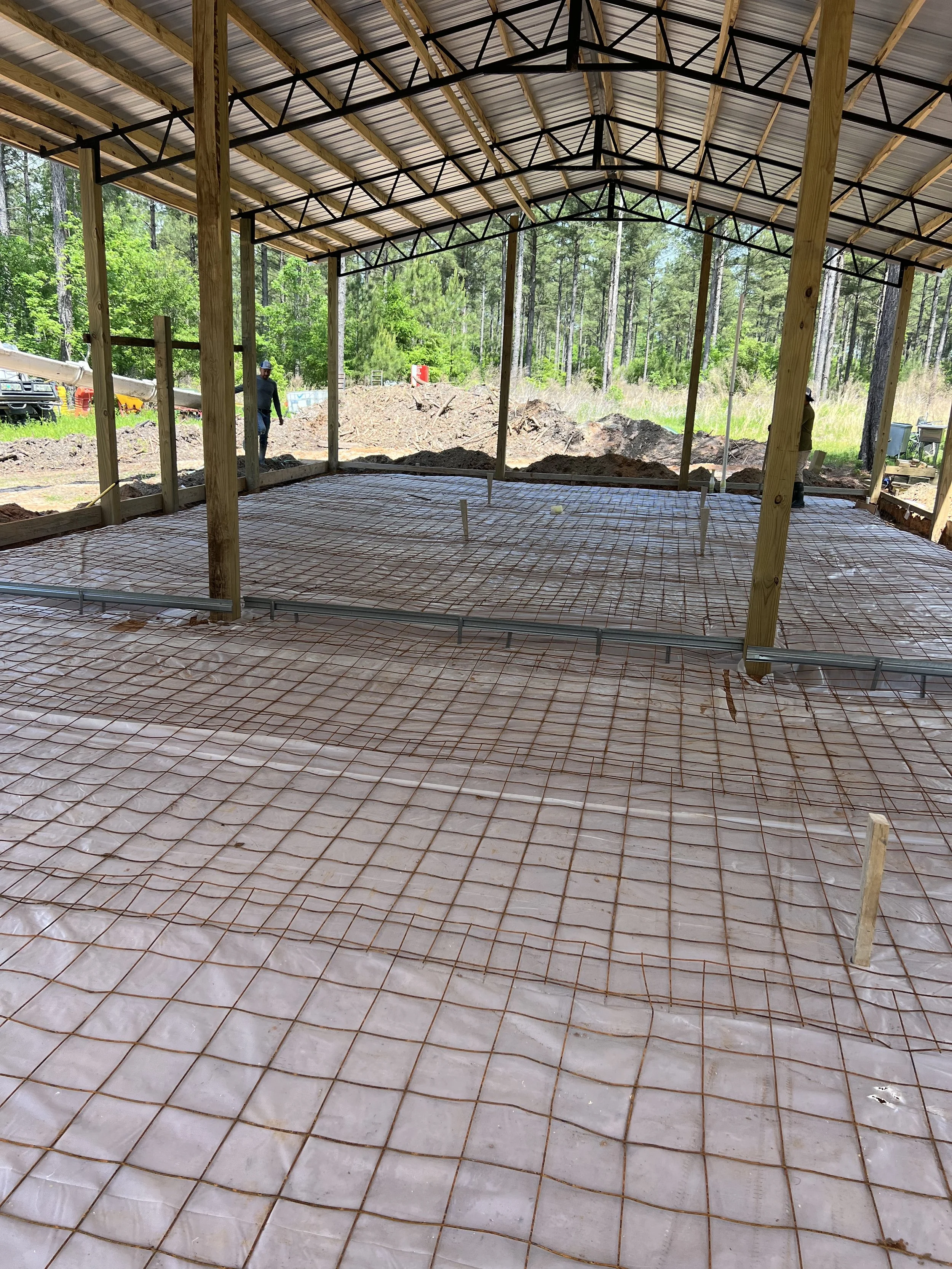 Construction site showing the foundation with rebar grid and wooden support beams for a building, with a metal roof and surrounded by trees.