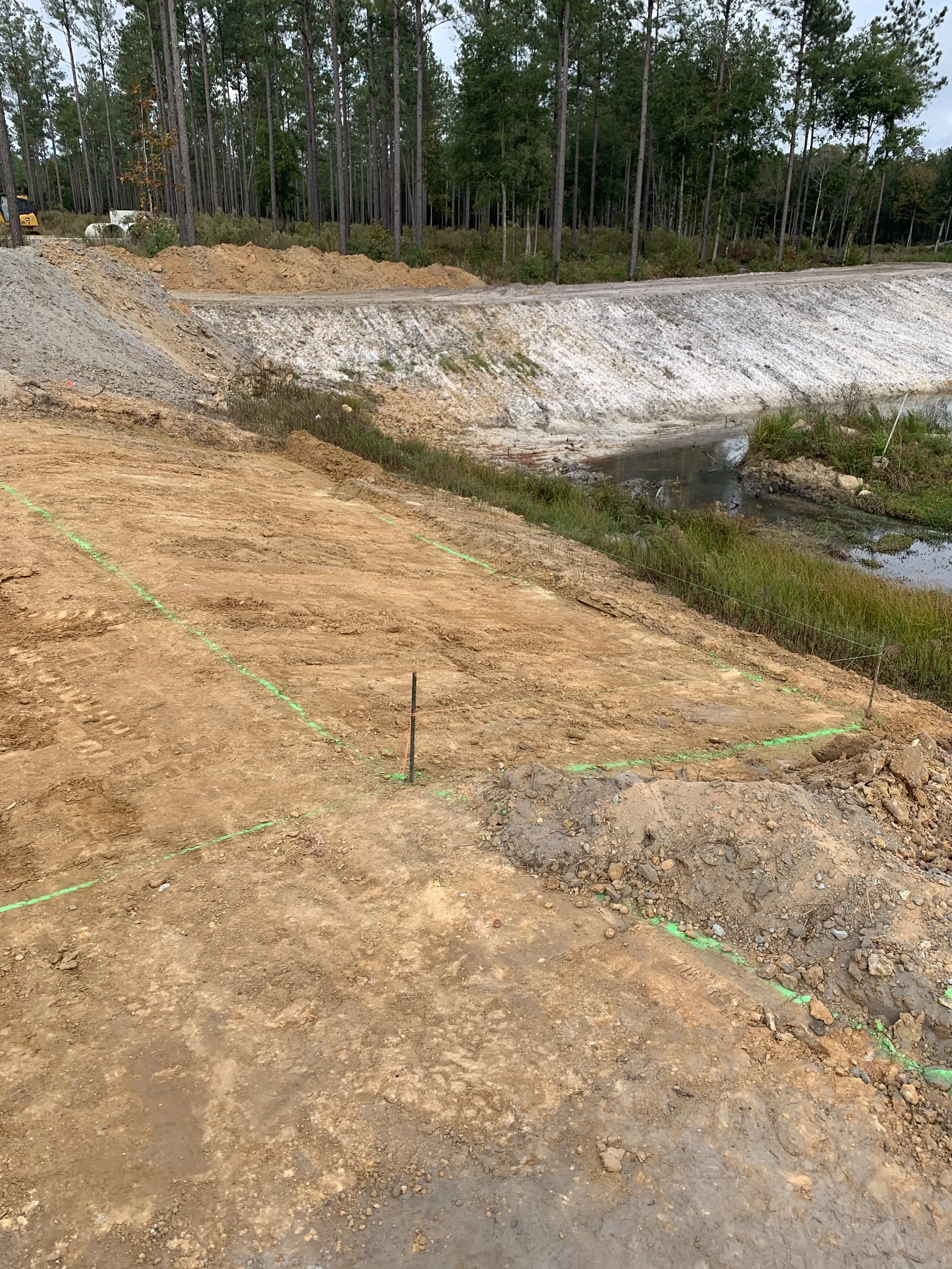 Construction site with marked excavation area, dirt, and construction markers, with a backdrop of trees and a small pond or water feature.