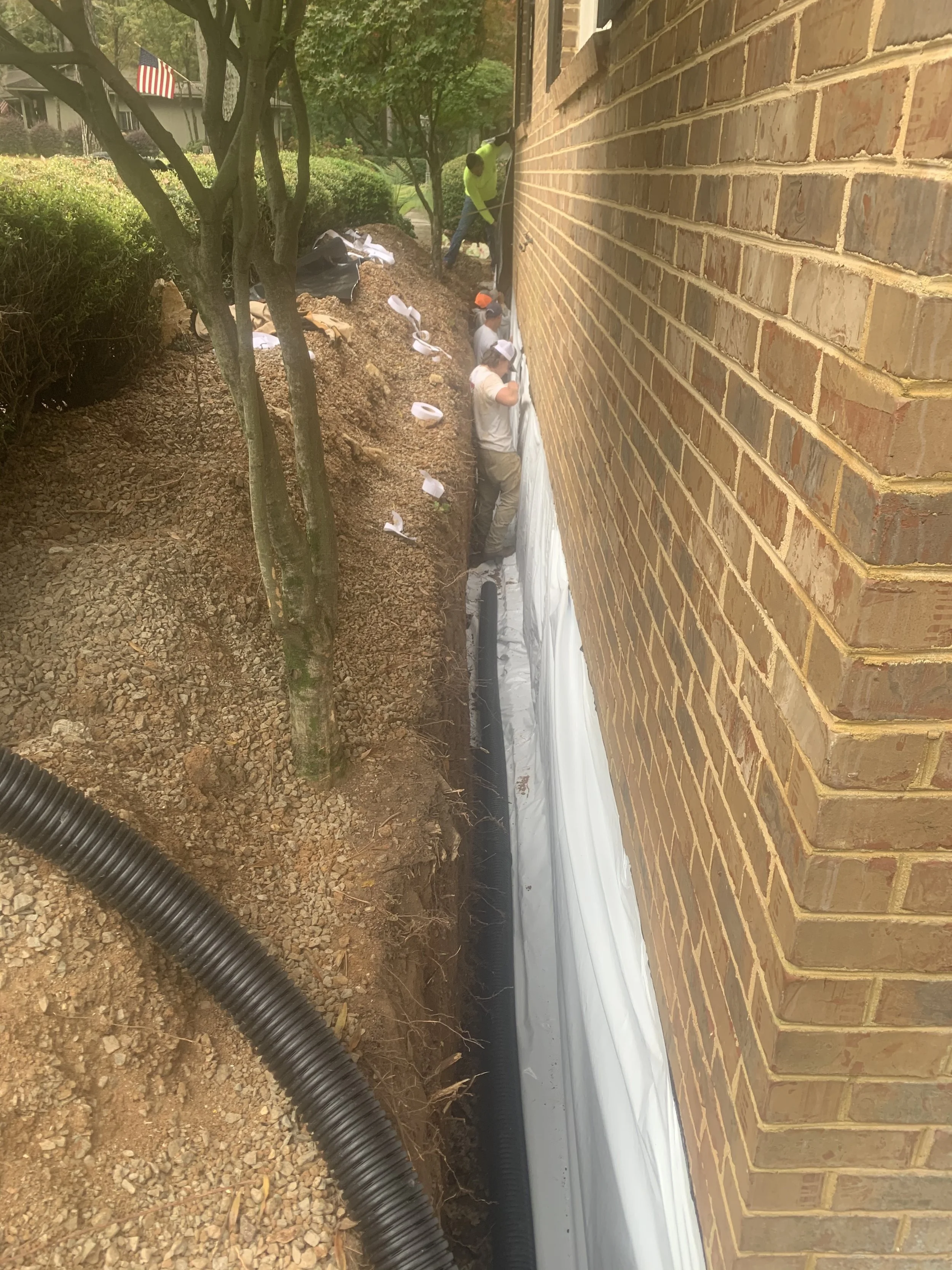 Workers installing a white vapor barrier along the foundation of a brick house. Some workers are on a ladder, while others work on the ground next to a trench filled with dirt and black drainage pipes. There are trees and bushes nearby, and an Americ