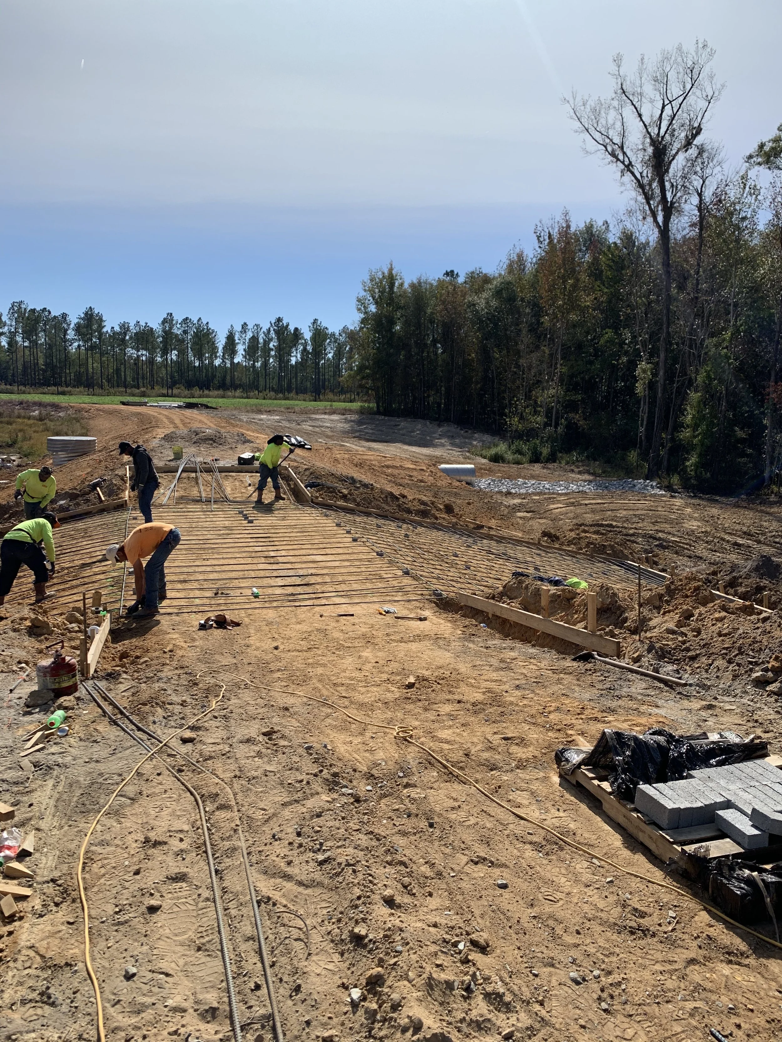 Construction workers laying down a concrete roadway at a construction site, with trees and a clear sky in the background.