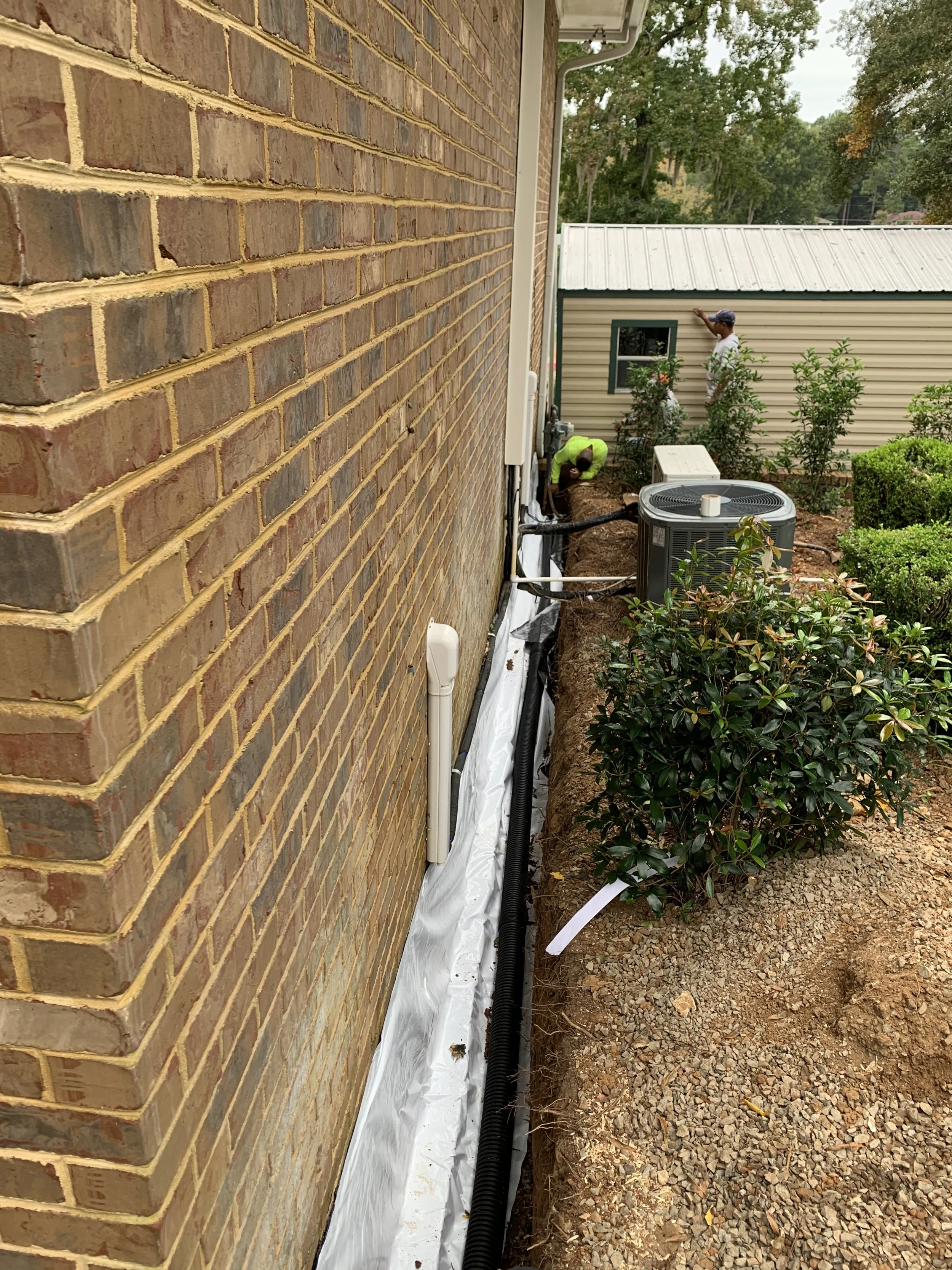Two workers are installing or repairing a drainage or utility system along the side of a brick house. One worker is kneeling near a black pipe, while the other is standing near the small window of a beige building or shed in the background, surrounde