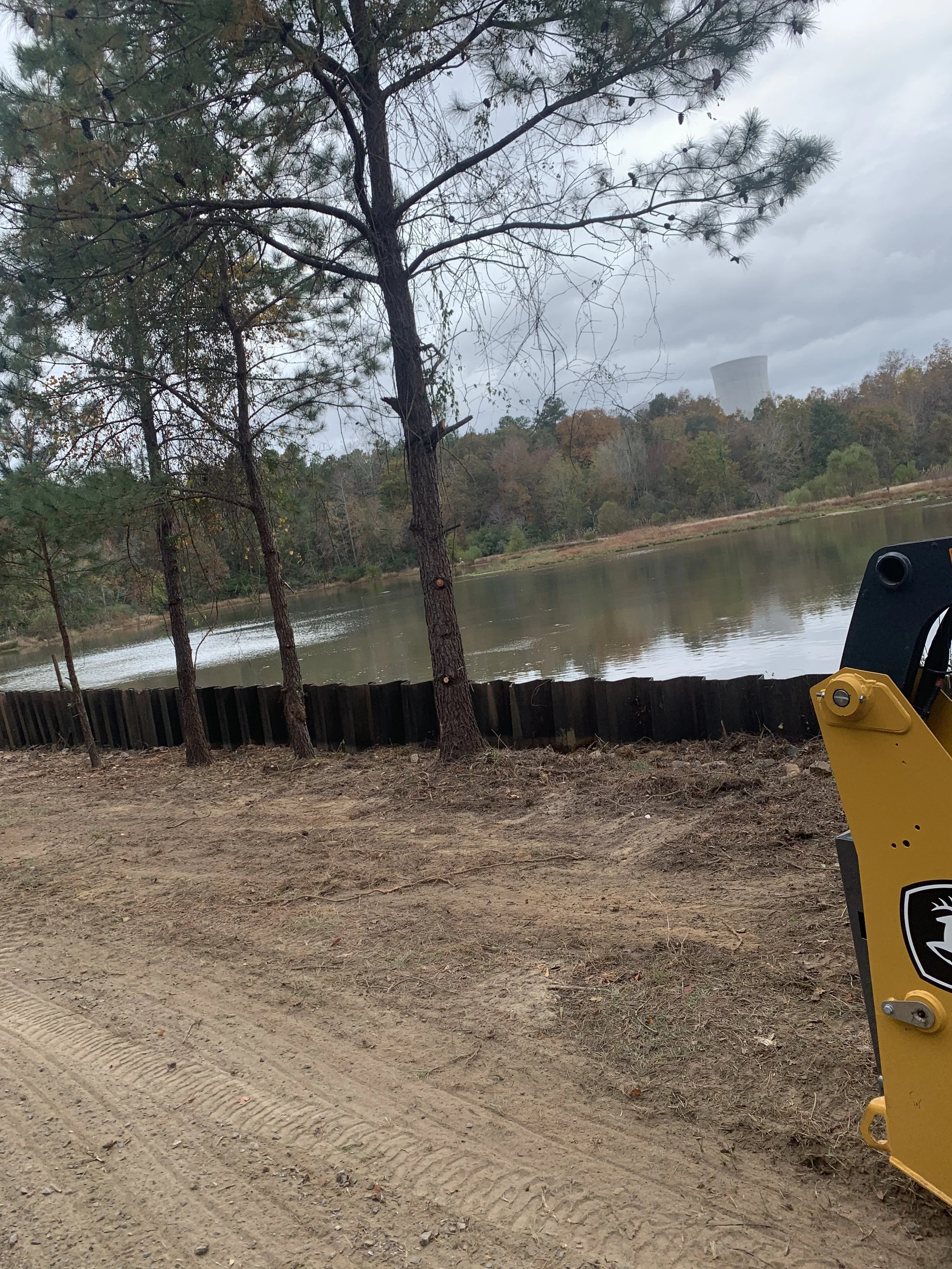 A natural landscape with a lake partly surrounded by trees, cloudy sky, and a distant industrial cooling tower, with part of construction equipment in the foreground.