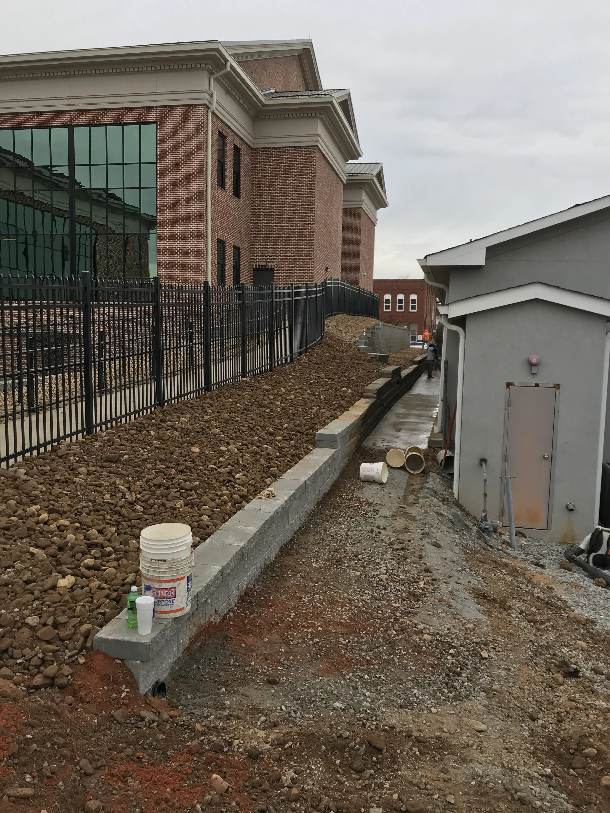 Construction site with a newly built cinder block wall, soil, and construction materials, with a large brick building and a black metal fence in the background.