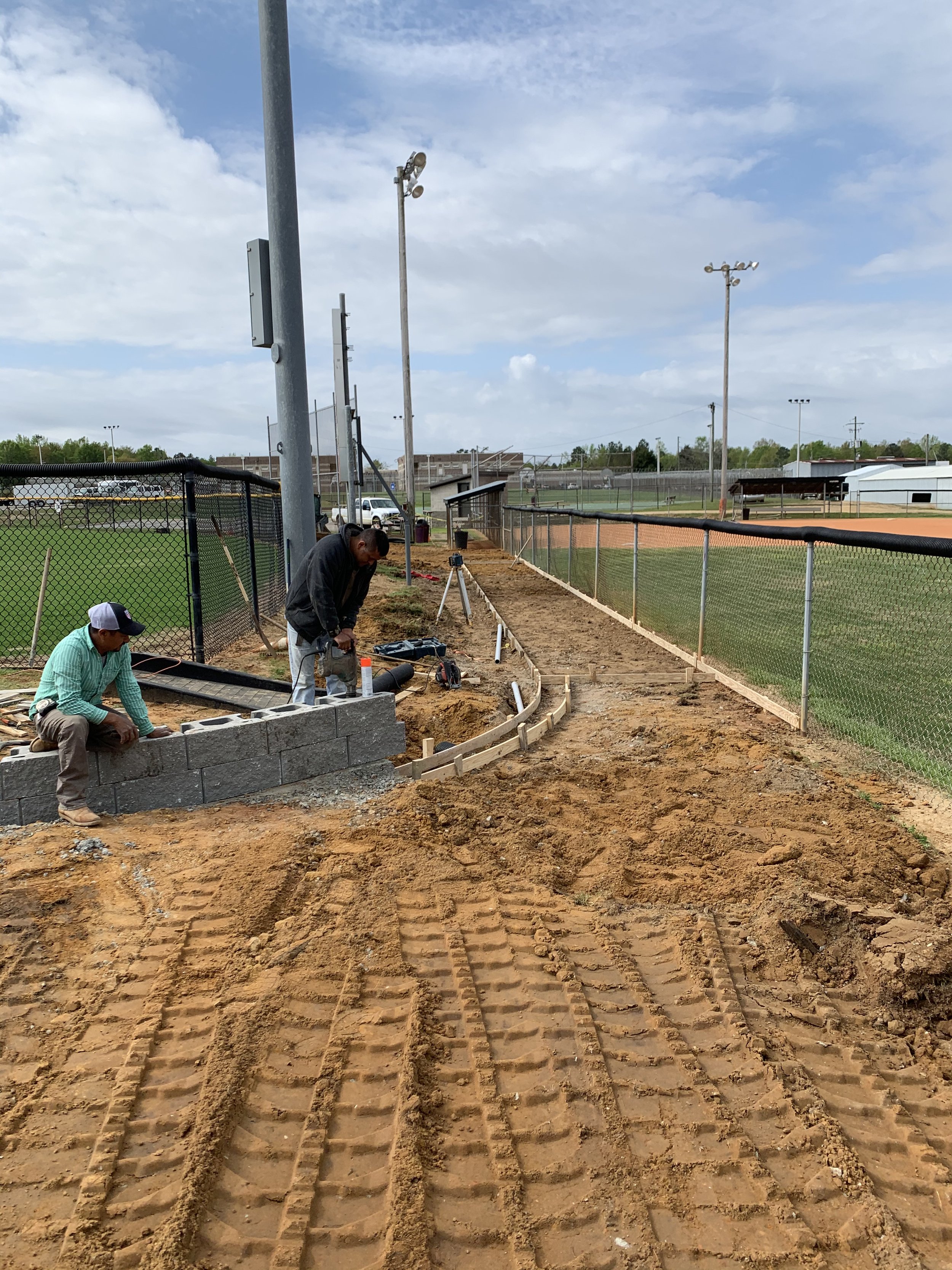 Construction workers build a curved brick wall along a fence at a baseball field under cloudy sky.