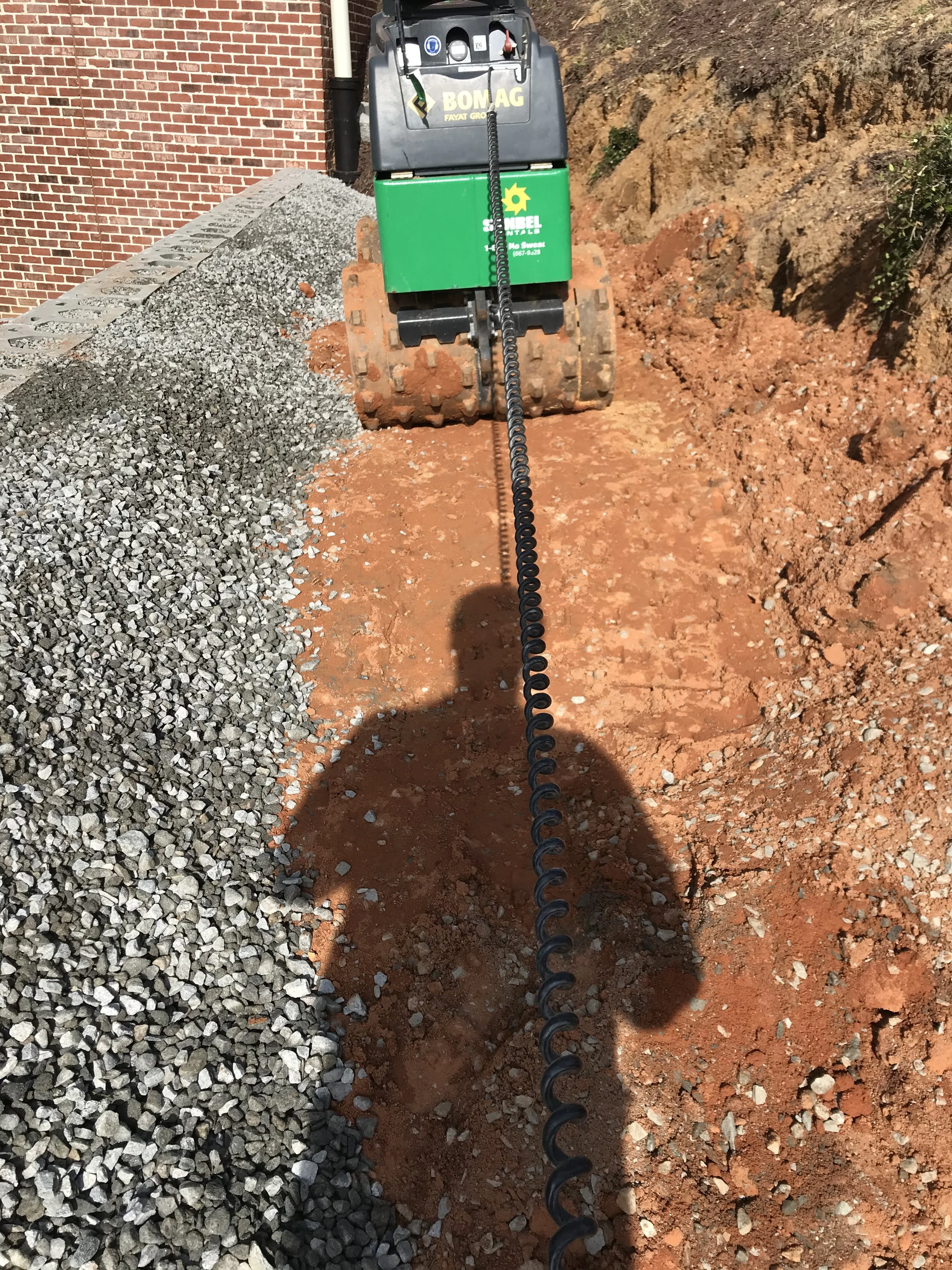 Small construction bulldozer with a chain attached, working on a dirt trench beside a brick building wall and a pile of gravel.