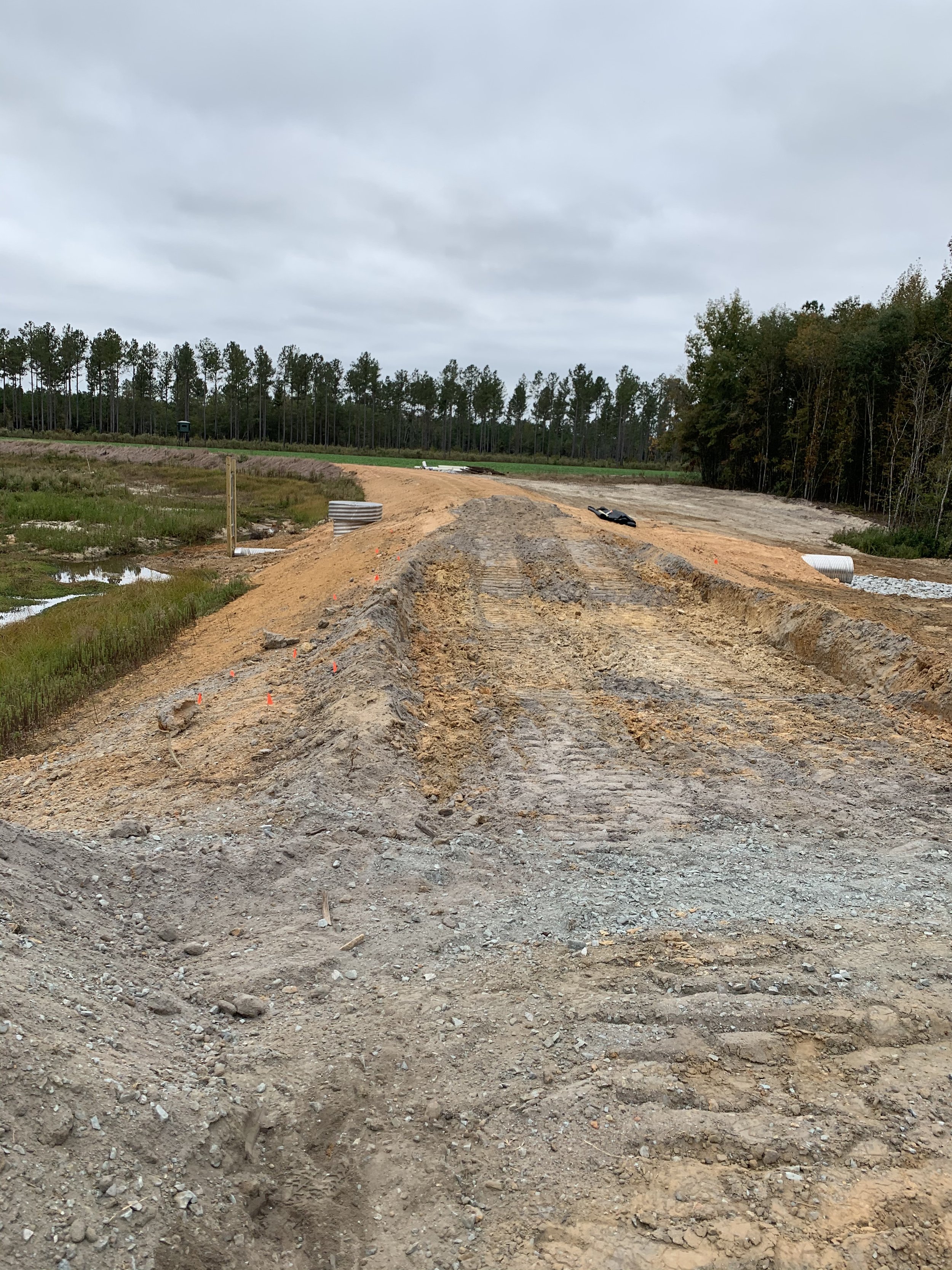 Construction site on an unfinished road with dirt, tire tracks, and construction materials, surrounded by trees and cloudy sky.
