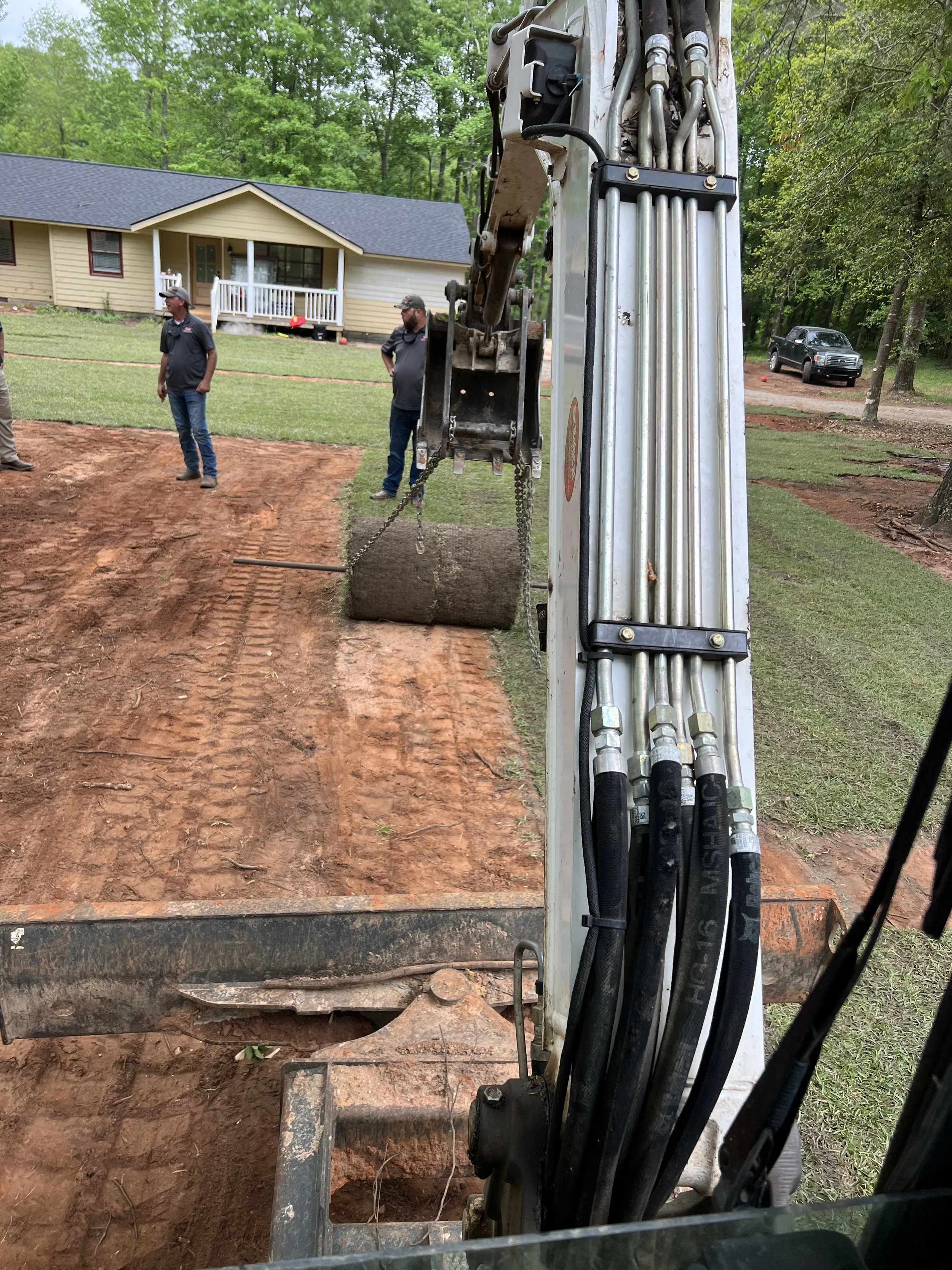 Construction workers and a mini excavator on a residential backyard renovation site, with a house and cars in the background.