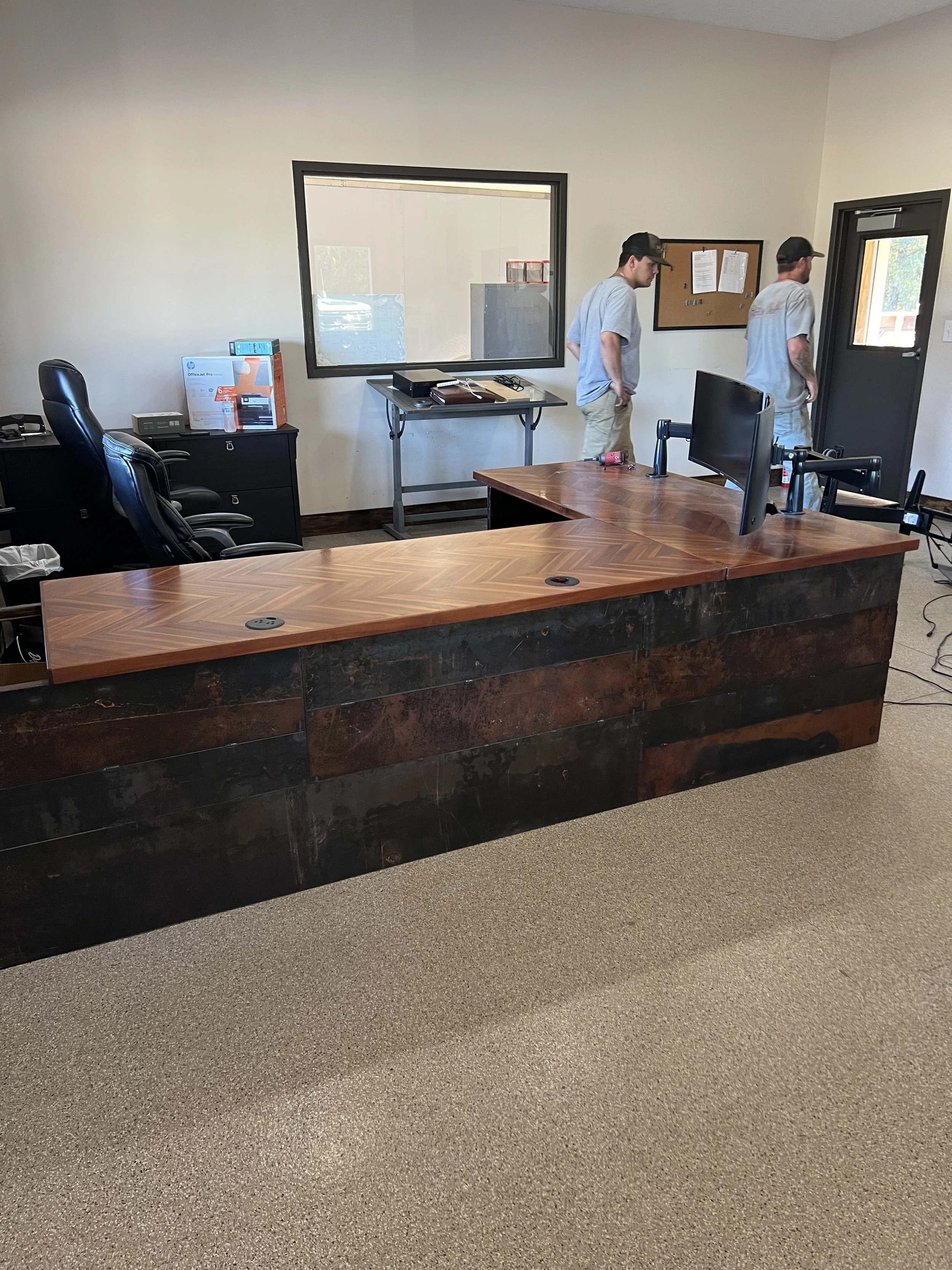 An office with a large wooden desk, several black office chairs, a computer monitor, and two men standing by a door, with a window on the wall behind.