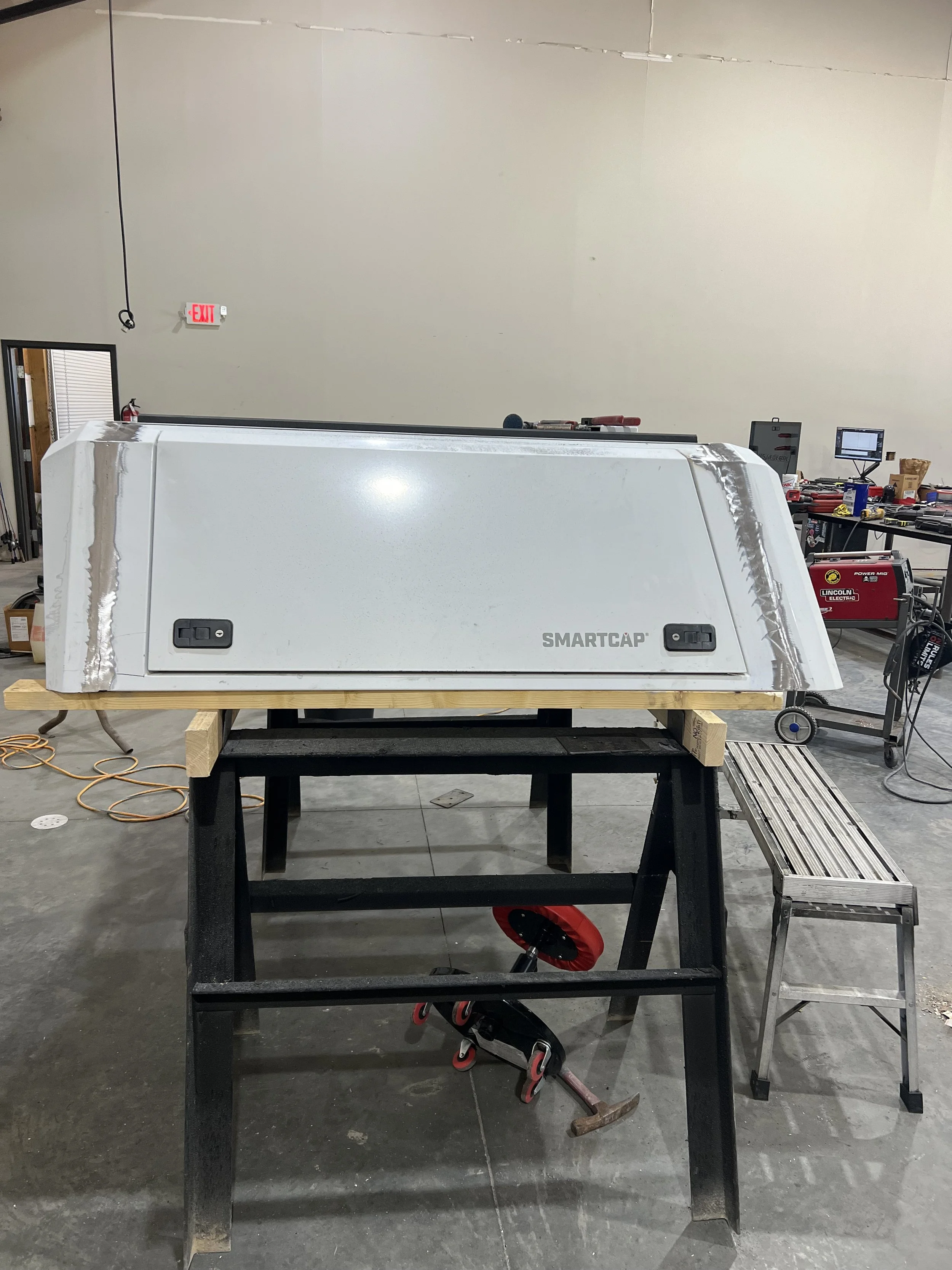 A large white truck bed topper labeled SMARTCAP on a black work table in a workshop with tools and equipment in the background.