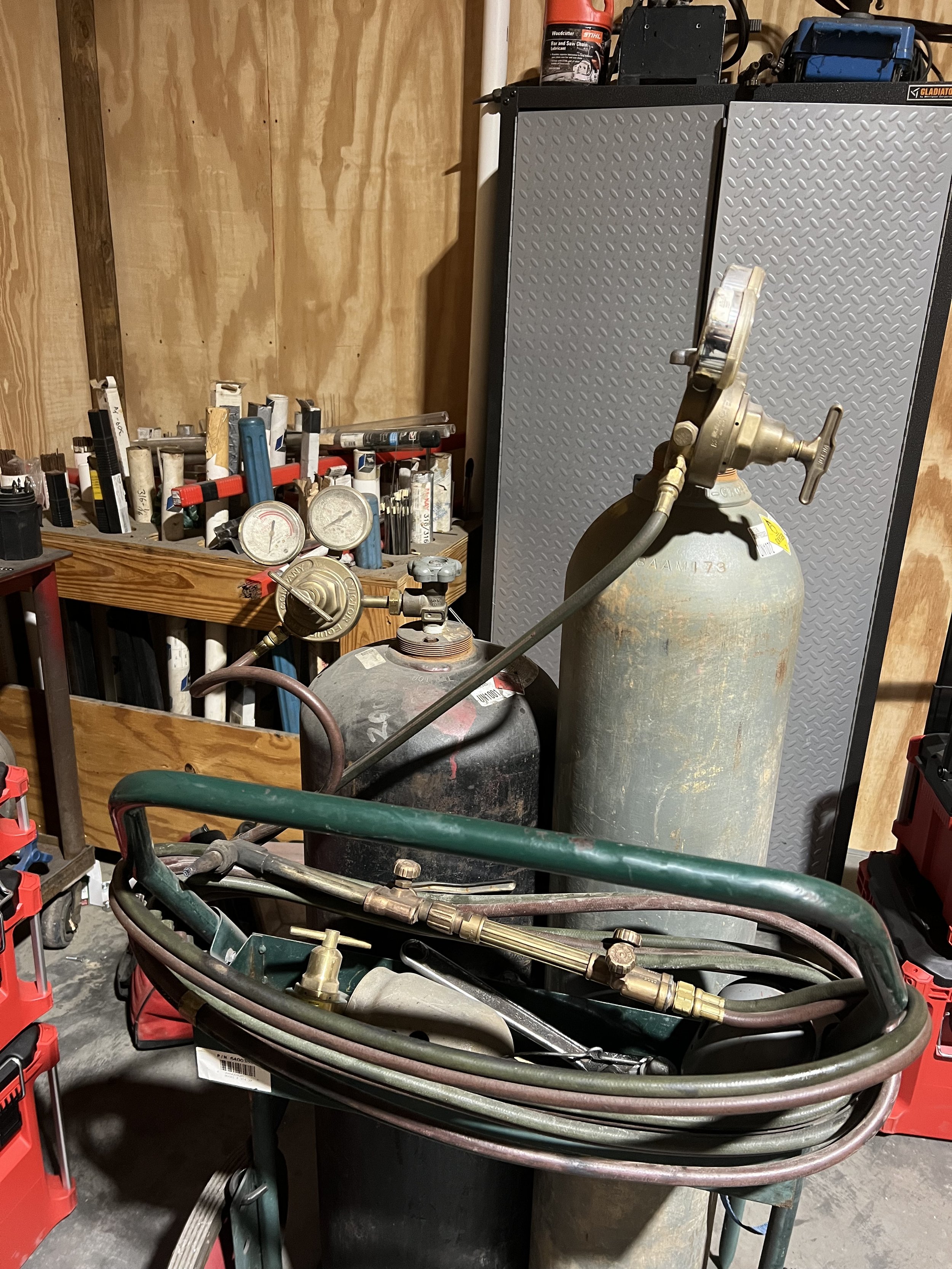 A workshop scene with two large gas cylinders, one black and one metallic, connected by hoses and fittings. In the background, work tools and gauges are visible along with a metal storage cabinet and a shelving unit filled with various small tools an