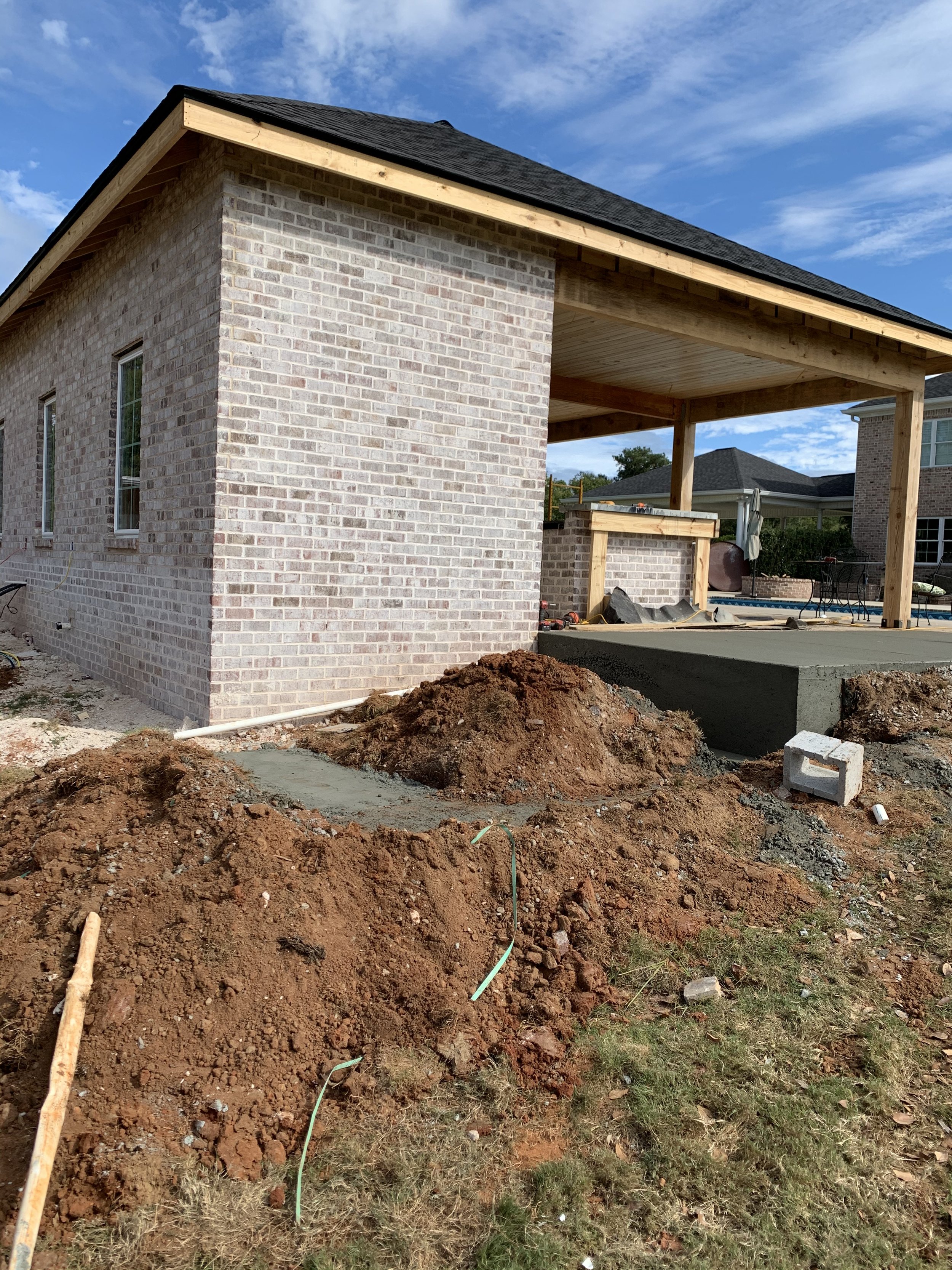 Construction site of a house with a brick wall, a porch with wooden beams, and a freshly poured concrete slab. Piles of dirt and construction materials are in the foreground, under a partly cloudy sky.