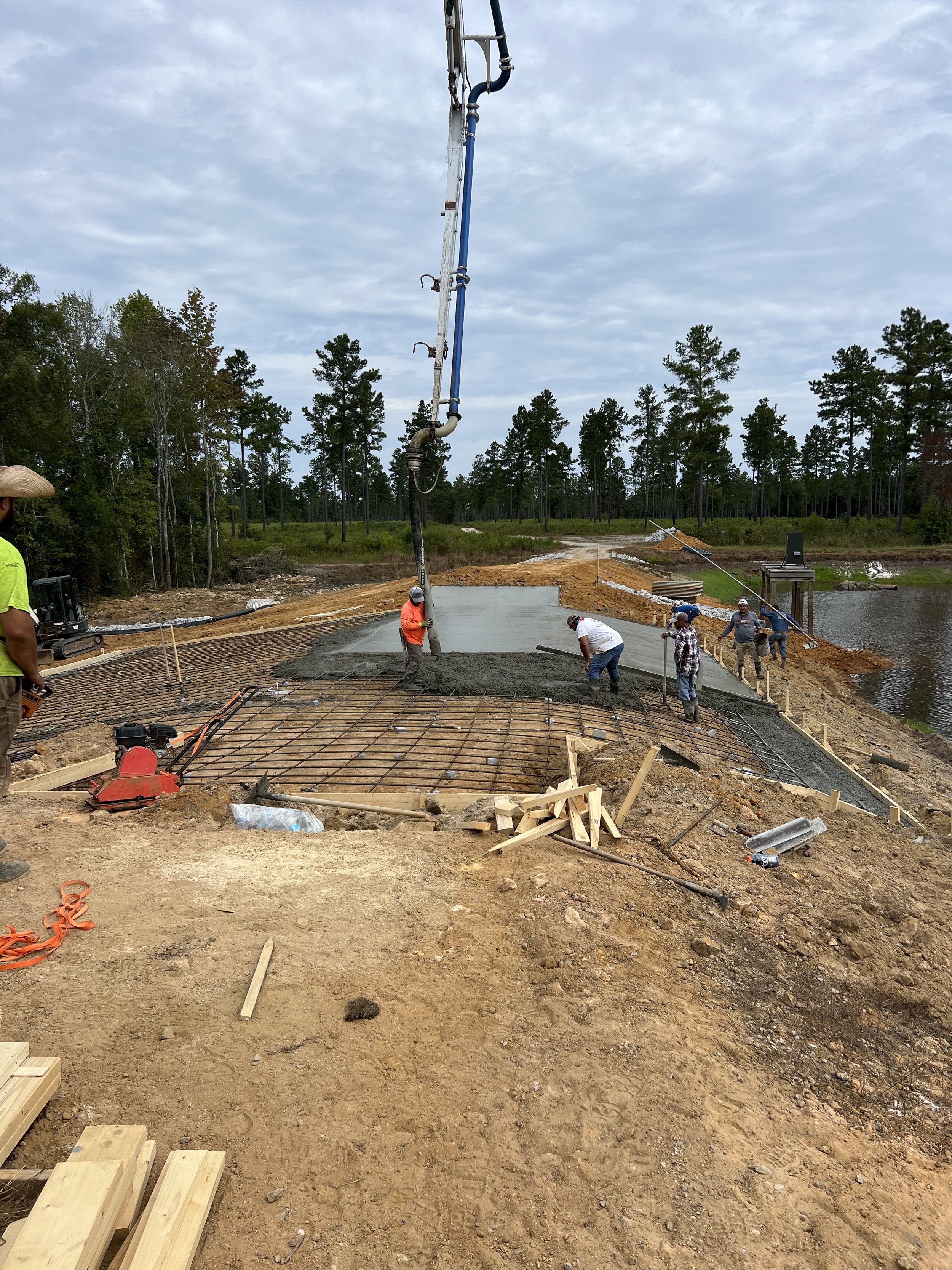 Construction workers pouring concrete on a foundation, with a reinforced steel grid in place, near a body of water surrounded by trees.