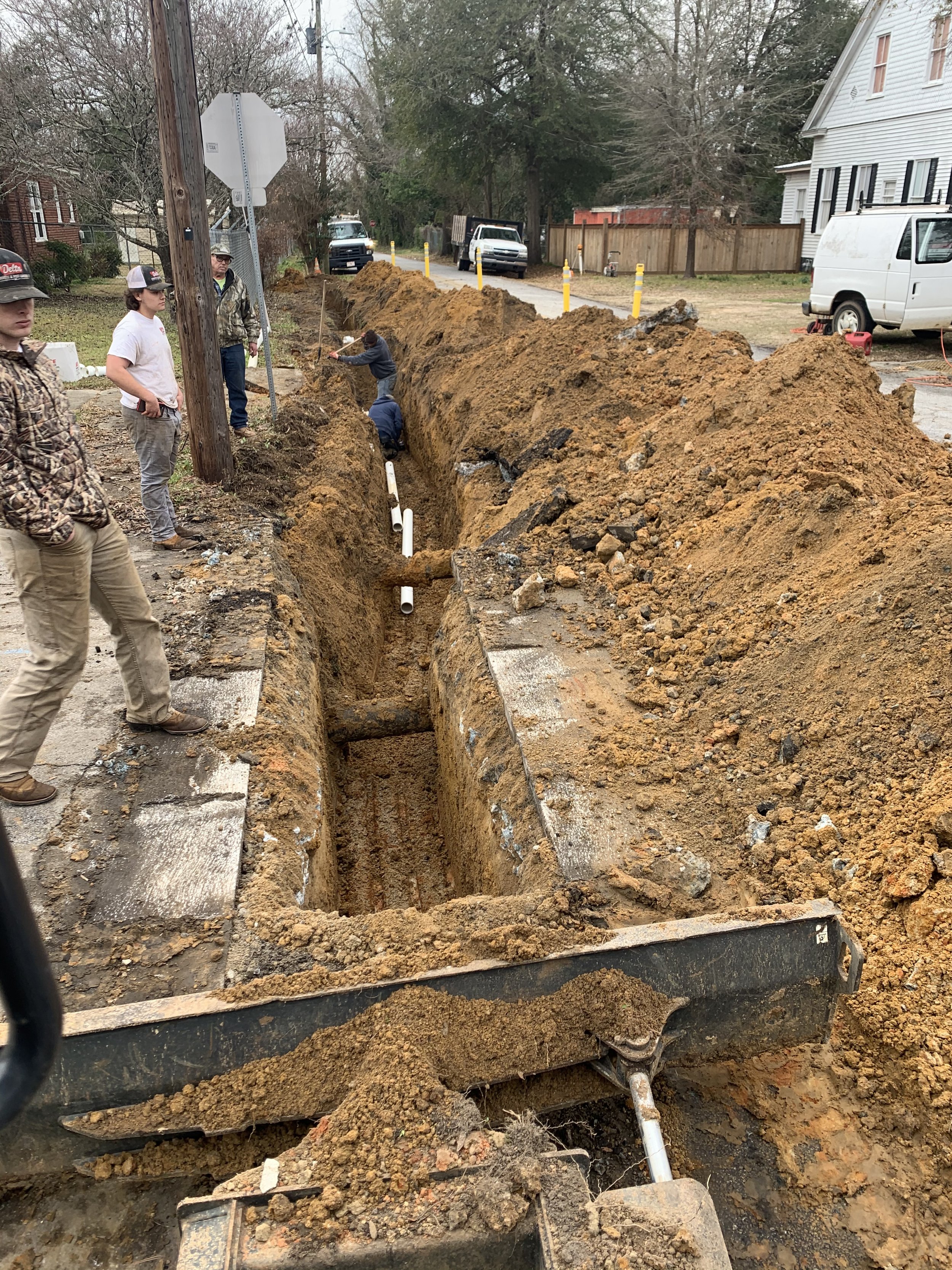 Construction workers digging a trench on a city street for underground pipes, with dirt piles along the road and houses in the background.