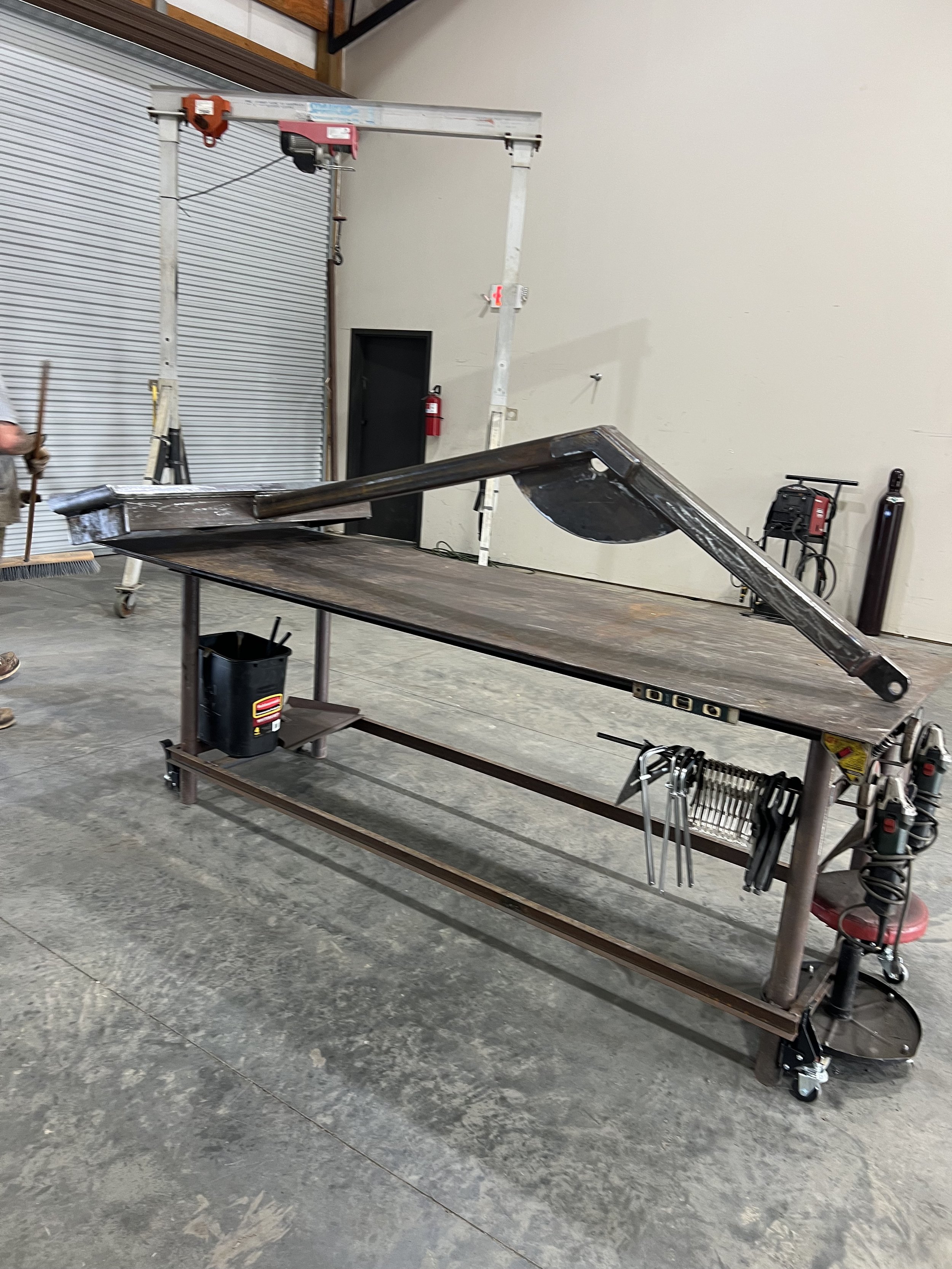 A large metal workbench with various tools hanging from its side, located in a workshop with concrete floors and a concrete wall with a closed metal roll-up door. There is a large cruciform go-kart chassis or framework on top of the workbench, likely