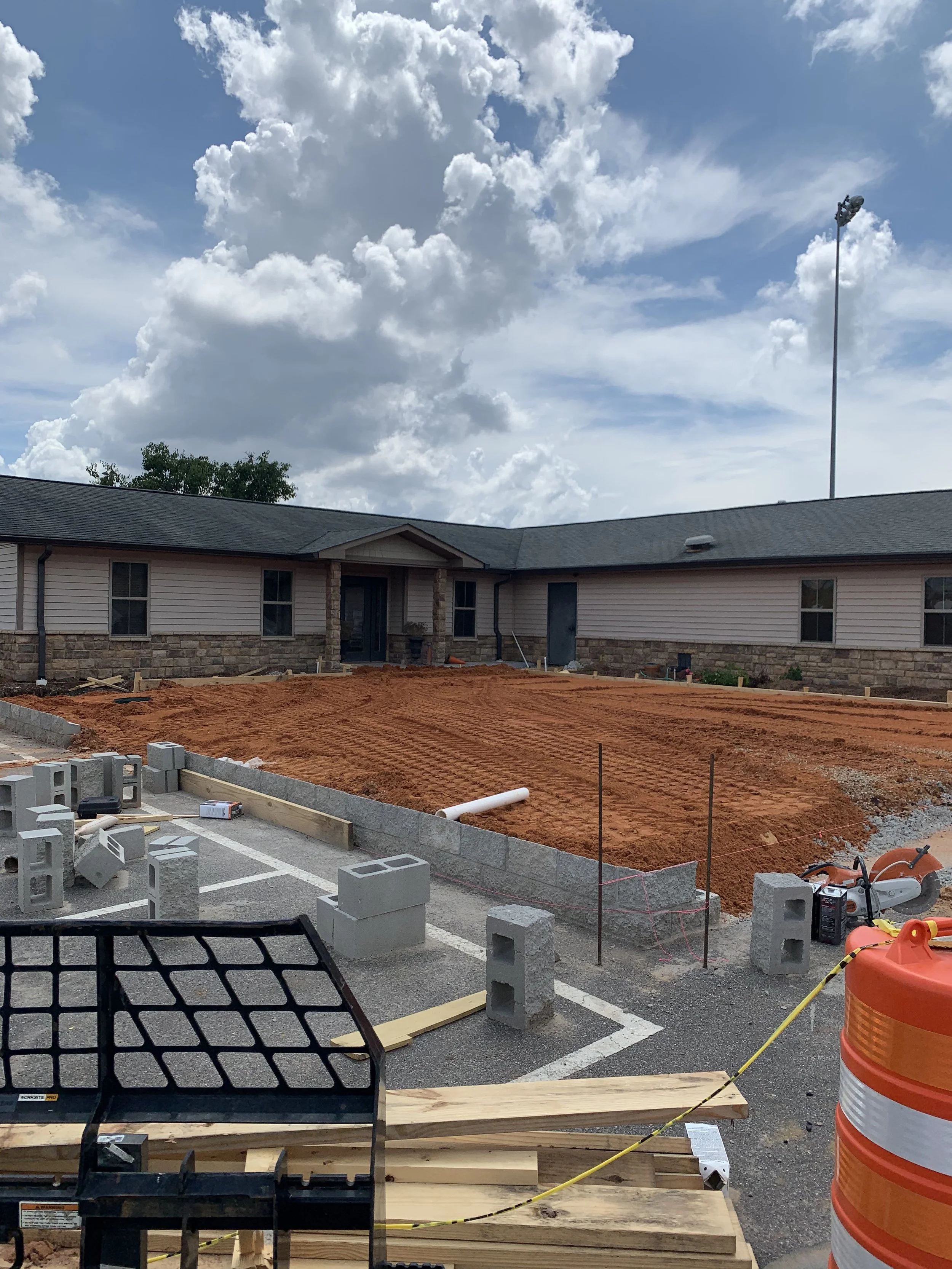 A construction site with a cleared dirt area in front of a building, cinder blocks, construction equipment, and a cloudy sky.