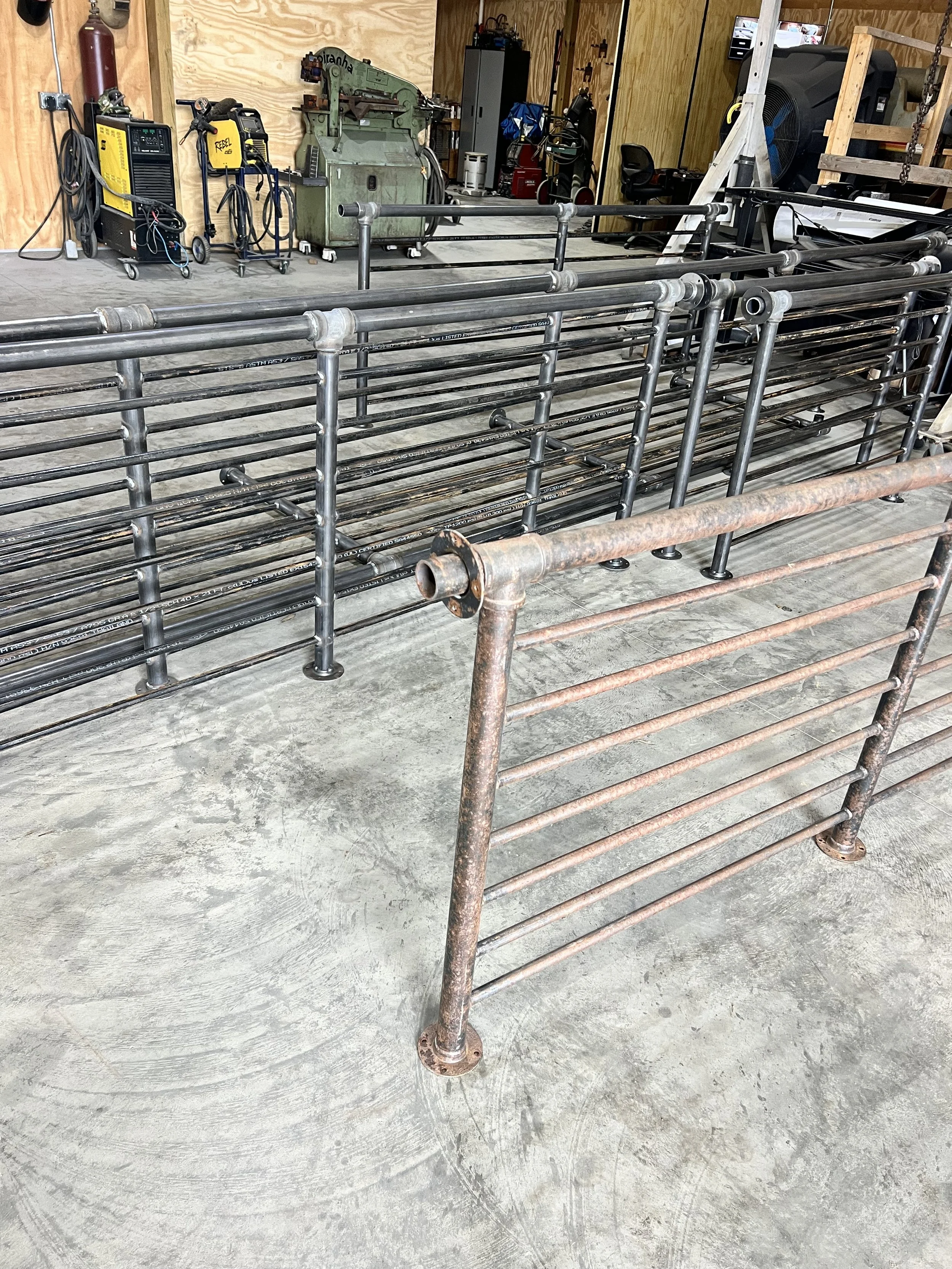 A variety of metal pipe fencing panels, some rusted and some new, stacked in a workshop or storage area with woodworking tools and equipment in the background.