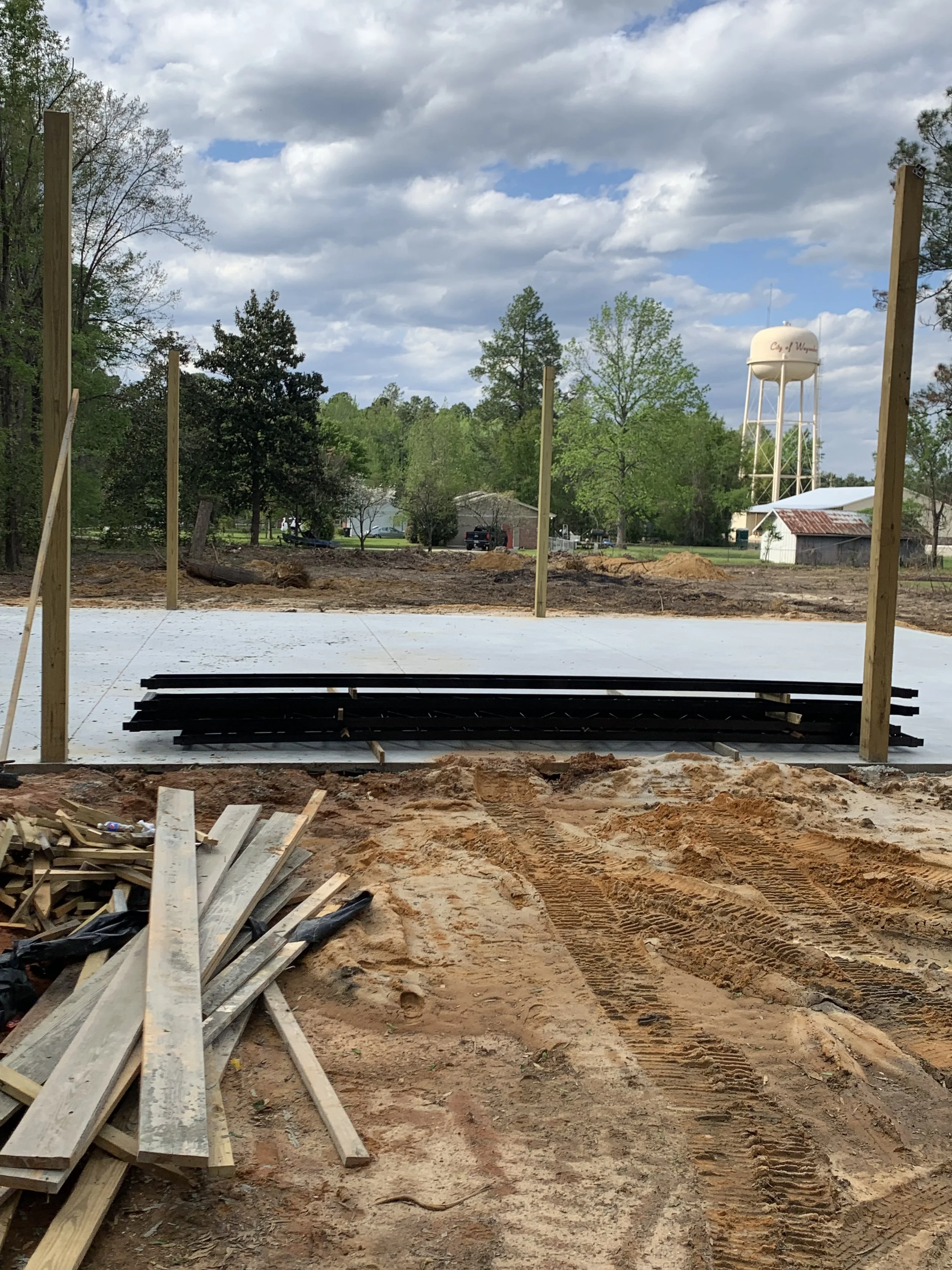 Construction site with a concrete foundation, building materials, and wooden posts. Trees, a water tower, and some buildings are visible in the background under a partly cloudy sky.