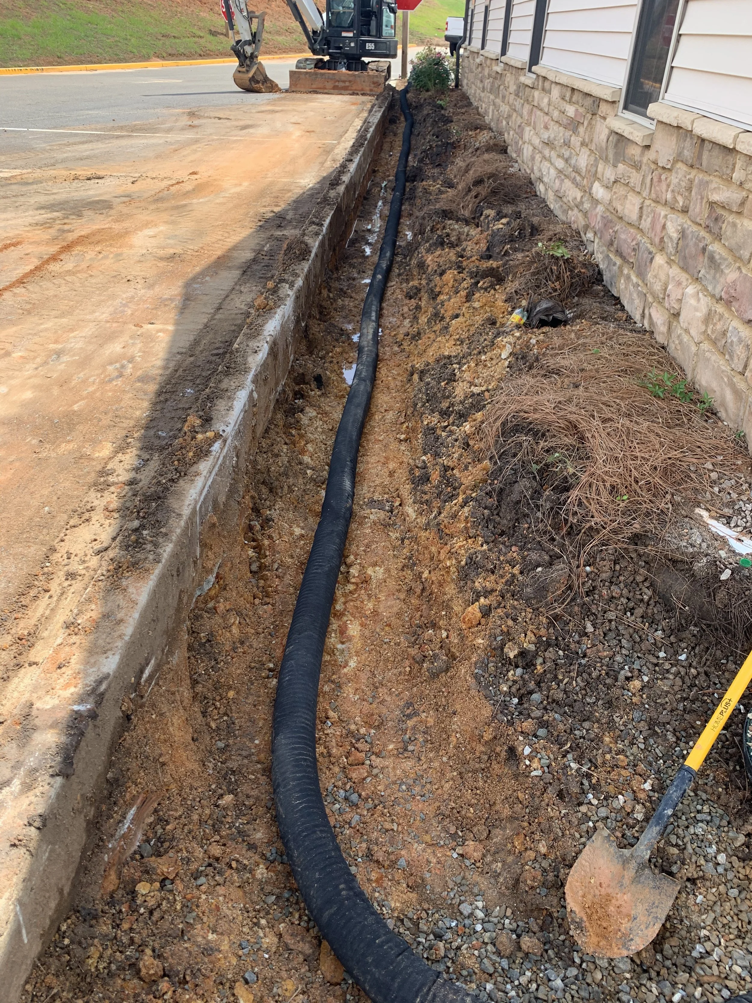 Construction site with a trench along a building wall, containing a black pipe, with a small excavator in the background.