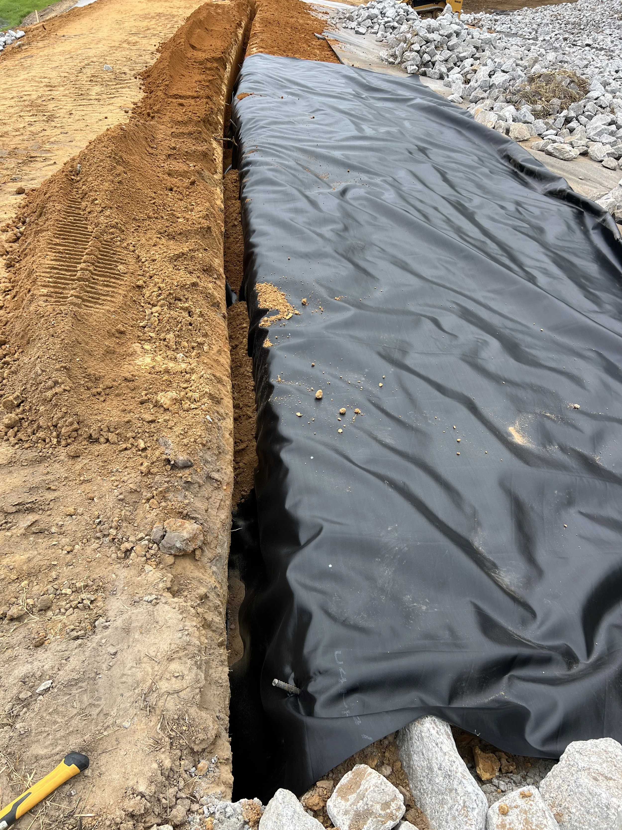 Construction site showing a trench covered with black plastic sheeting, with dirt and rocks nearby.