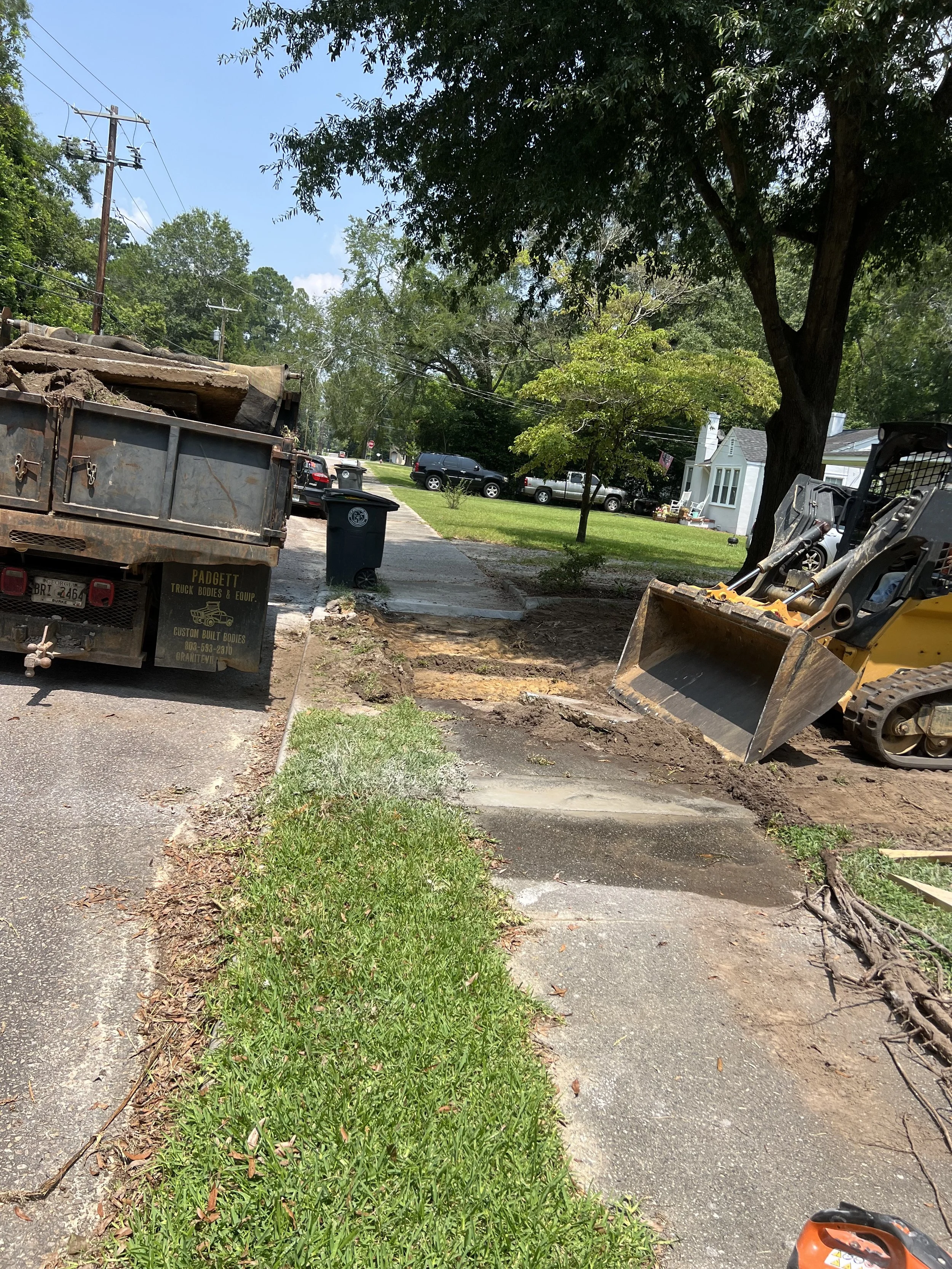Sidewalk and street under construction with a small excavator on the right and a dump truck on the left, trees, parked cars, and a blue trash bin in the background.