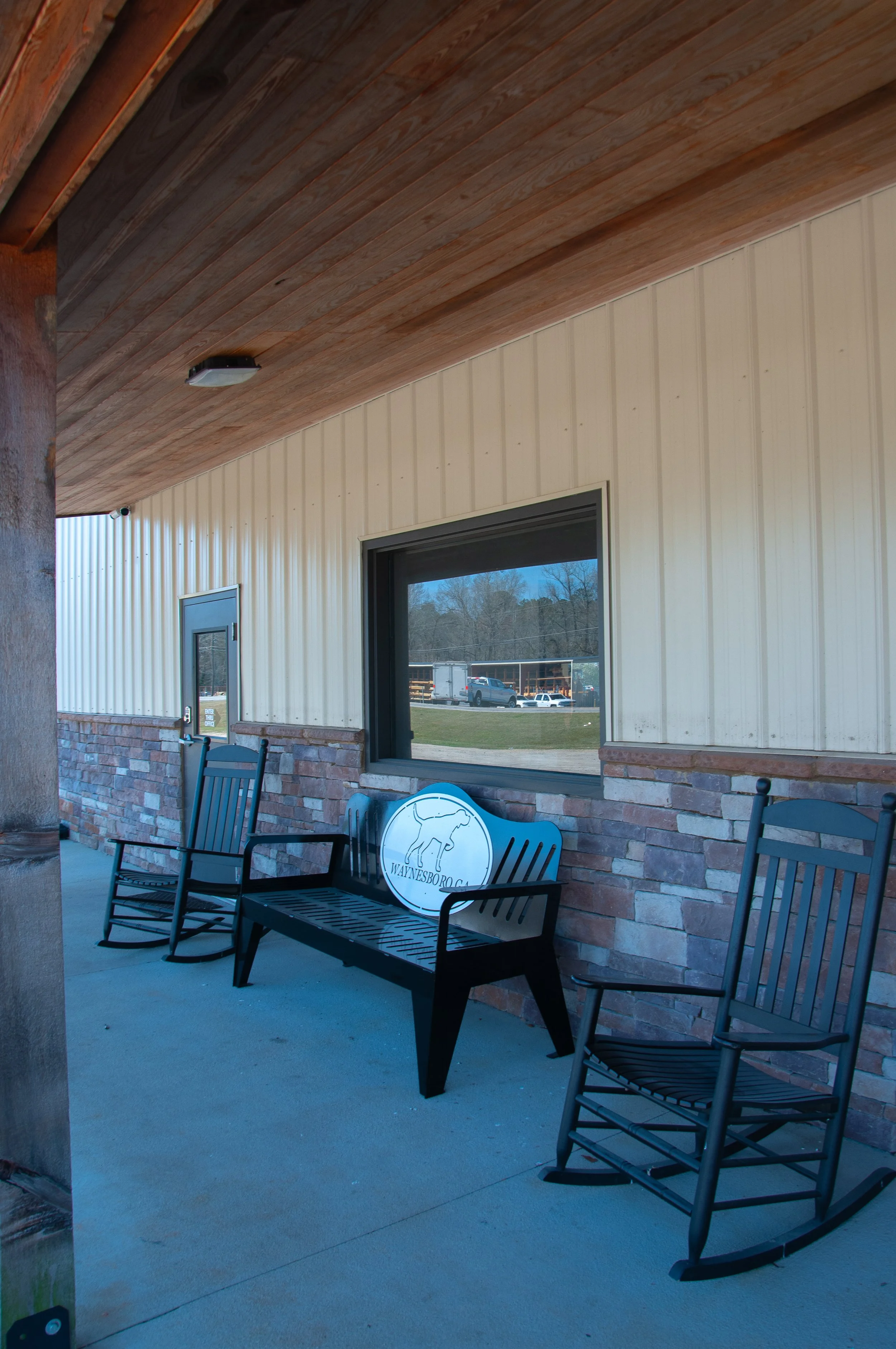 Outdoor seating area with three black rocking chairs and a black bench featuring a sign with a goat outline and the text 'WAYNESBORO, GA.' in front of a building with a beige metal wall and brick accent. A window reflects a parking lot and trees.