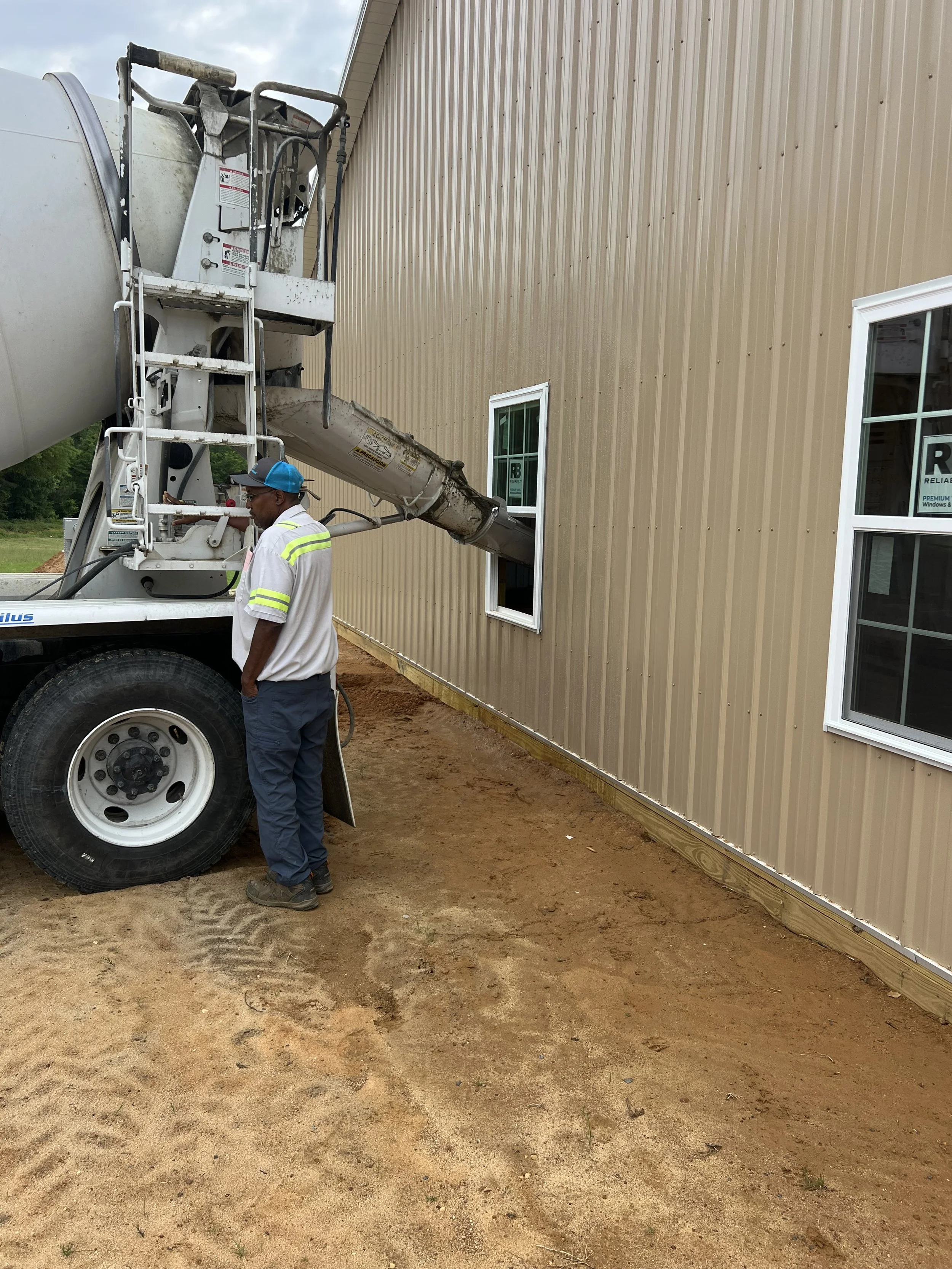 Construction worker in a white shirt and blue cap stands beside a cement mixer truck parked next to a beige building with white-framed windows. The cement chute extends into a window of the building.