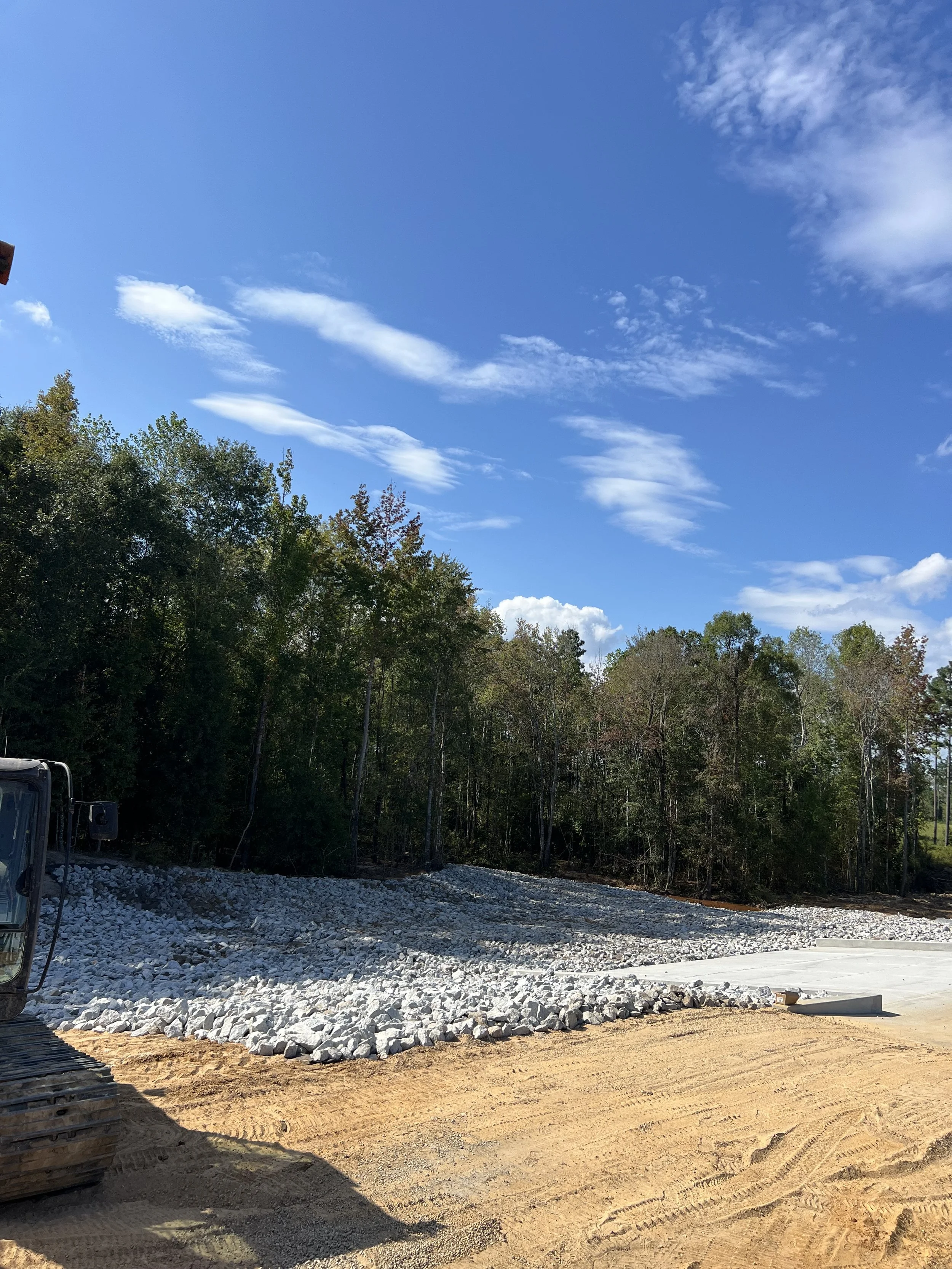 Construction site with gravel and dirt, trees in the background, and a blue sky with clouds.