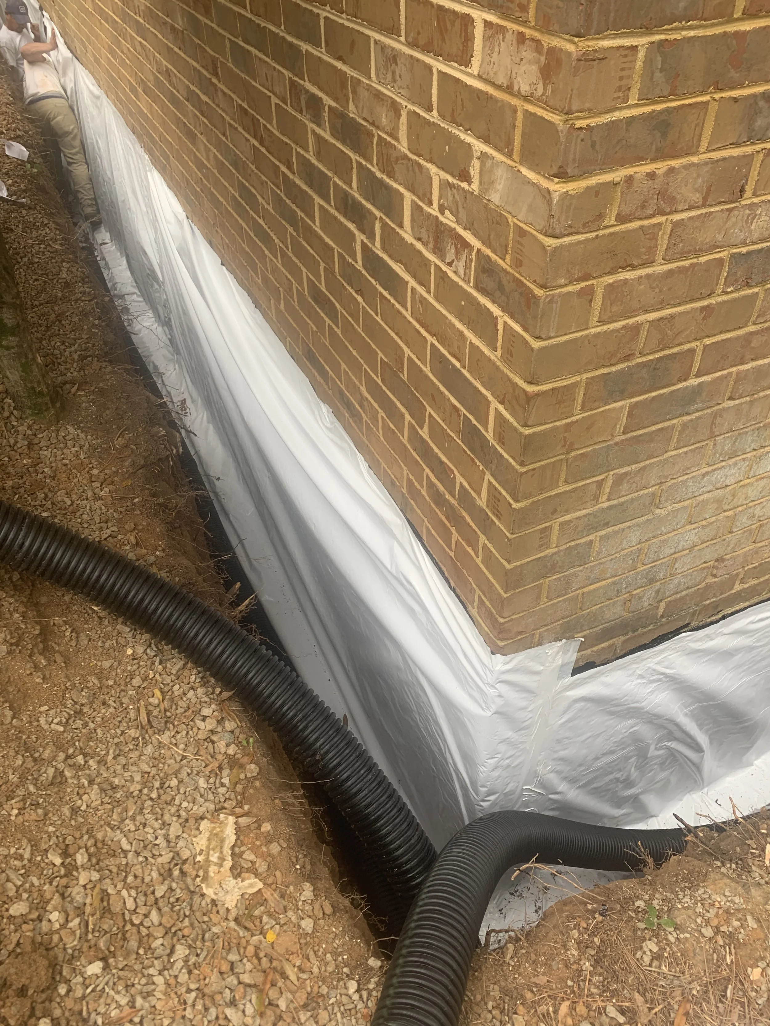 Foundation drainage system installation with a plastic liner and black drainage pipes next to a brick house wall.