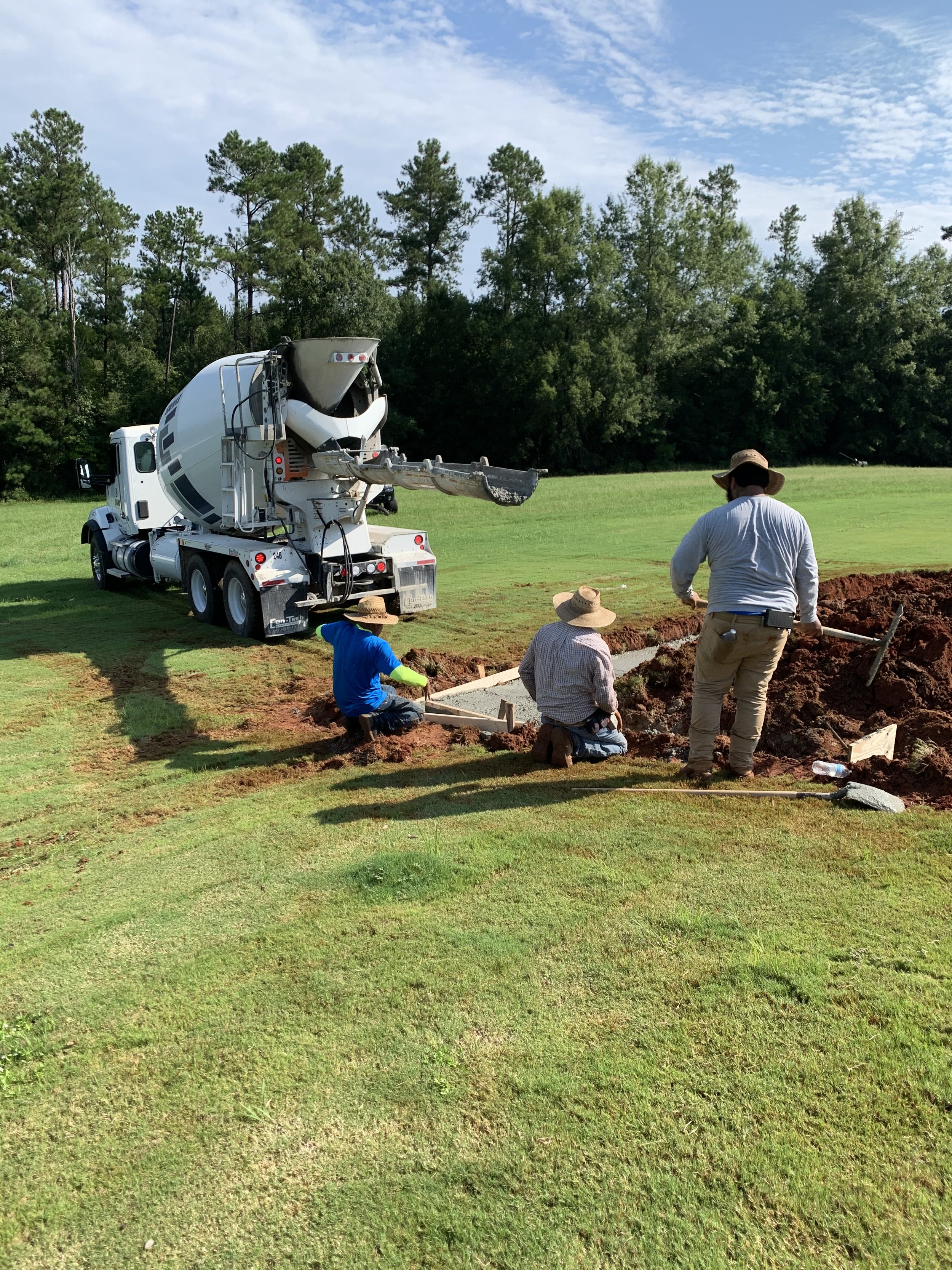 Workers pour concrete into a trench on a grassy field, with a concrete mixer truck nearby and trees in the background.