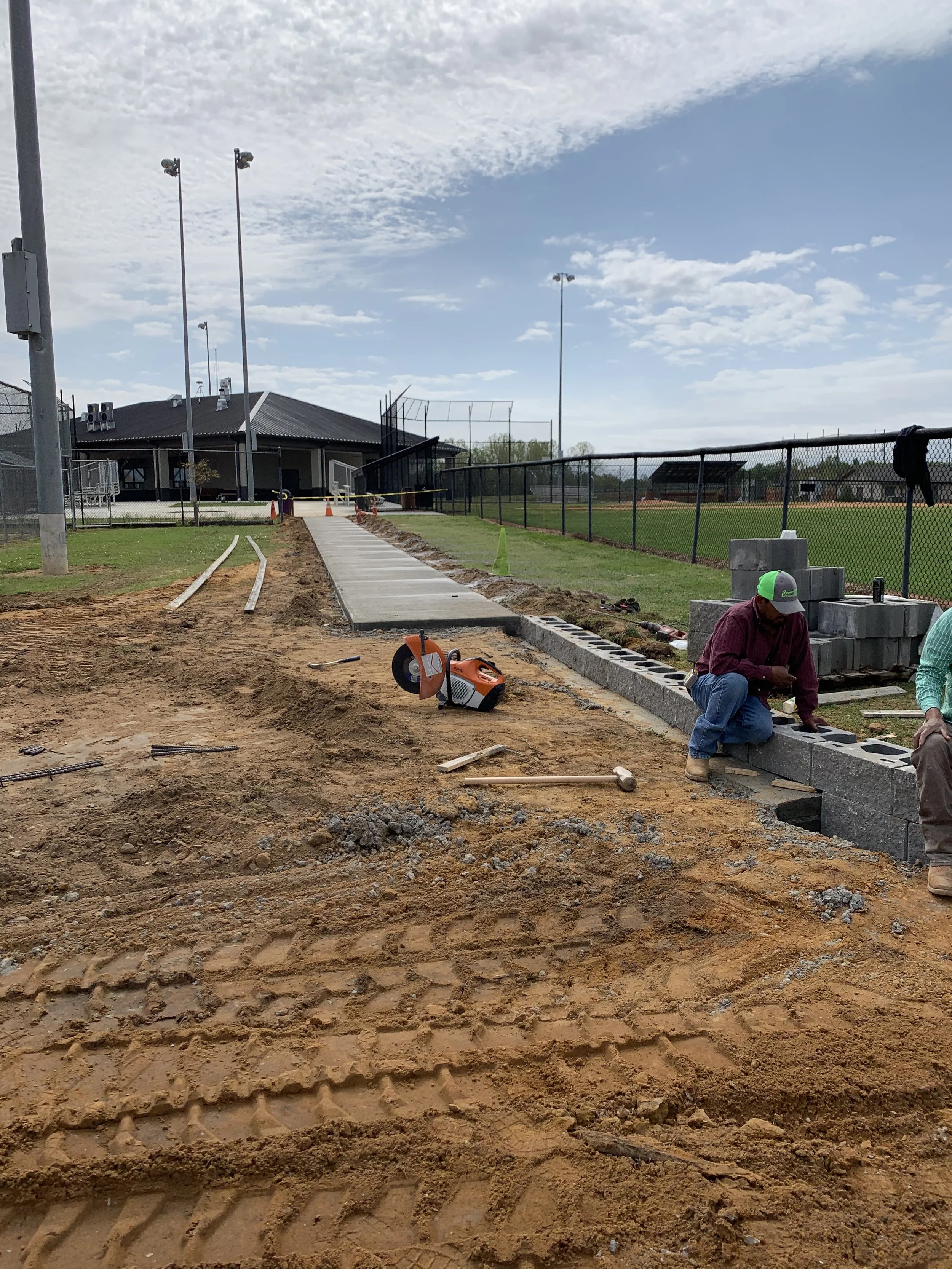 Construction workers laying concrete pavers along a baseball field fence with a chain-link fence, field, and baseball diamond in the background.