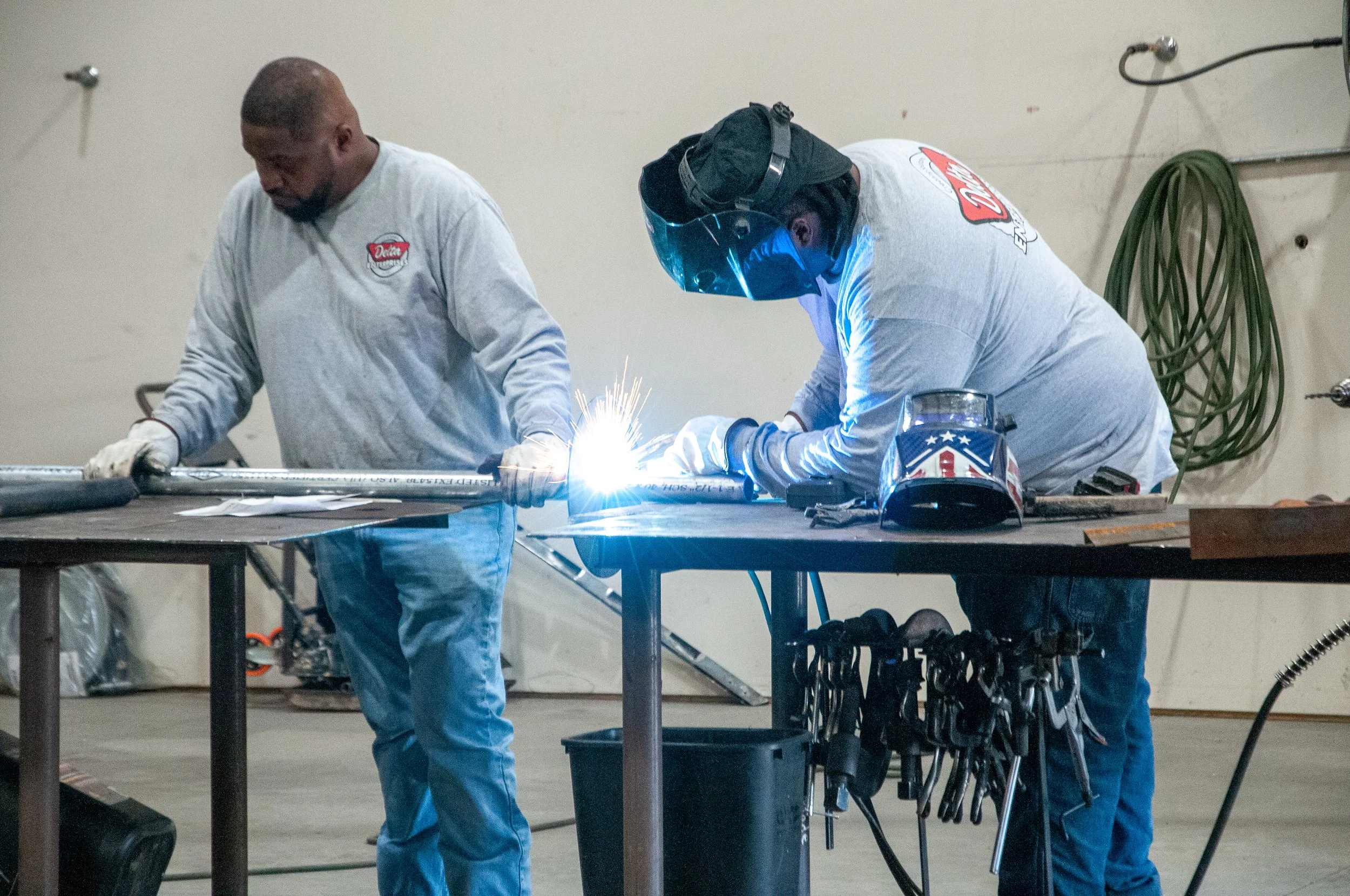 Two men welding a metal pipe in a workshop. One man is wearing a welding helmet, gloves, and a long-sleeve shirt, while the other man is watching. Welding sparks are visible.