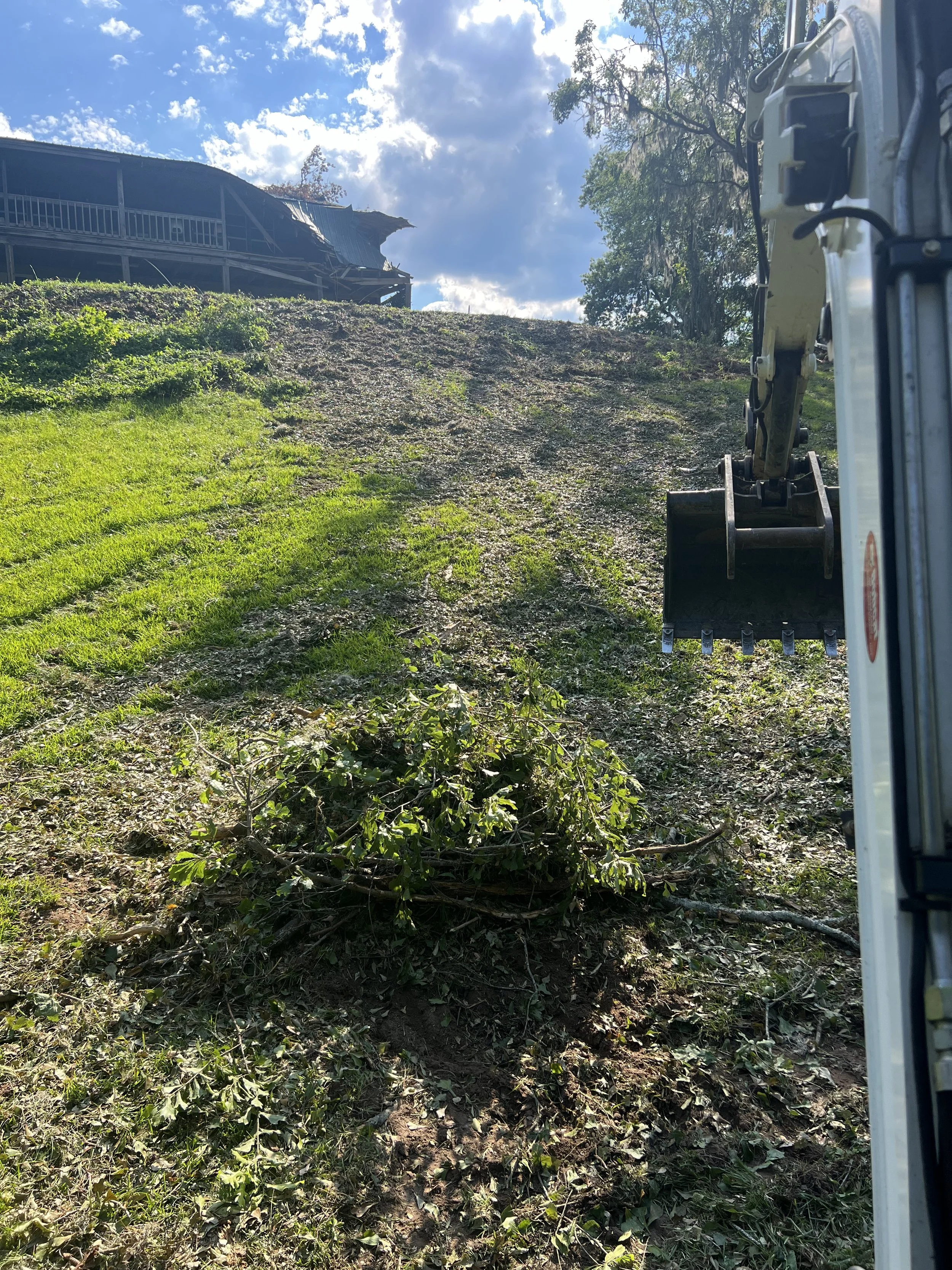 View from a construction vehicle showing a slope being cleared of small branches and leaves, with a damaged building in the background under a partly cloudy sky.
