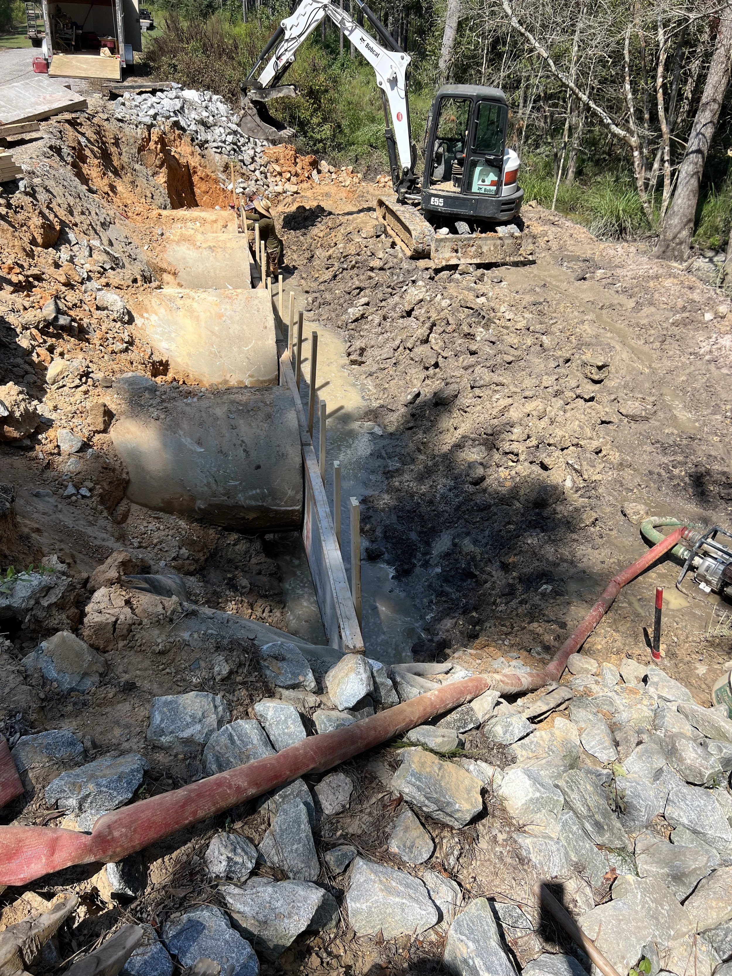 Construction site with an excavator digging a trench, with a worker in a straw hat inside the trench, surrounded by rocks and dirt.