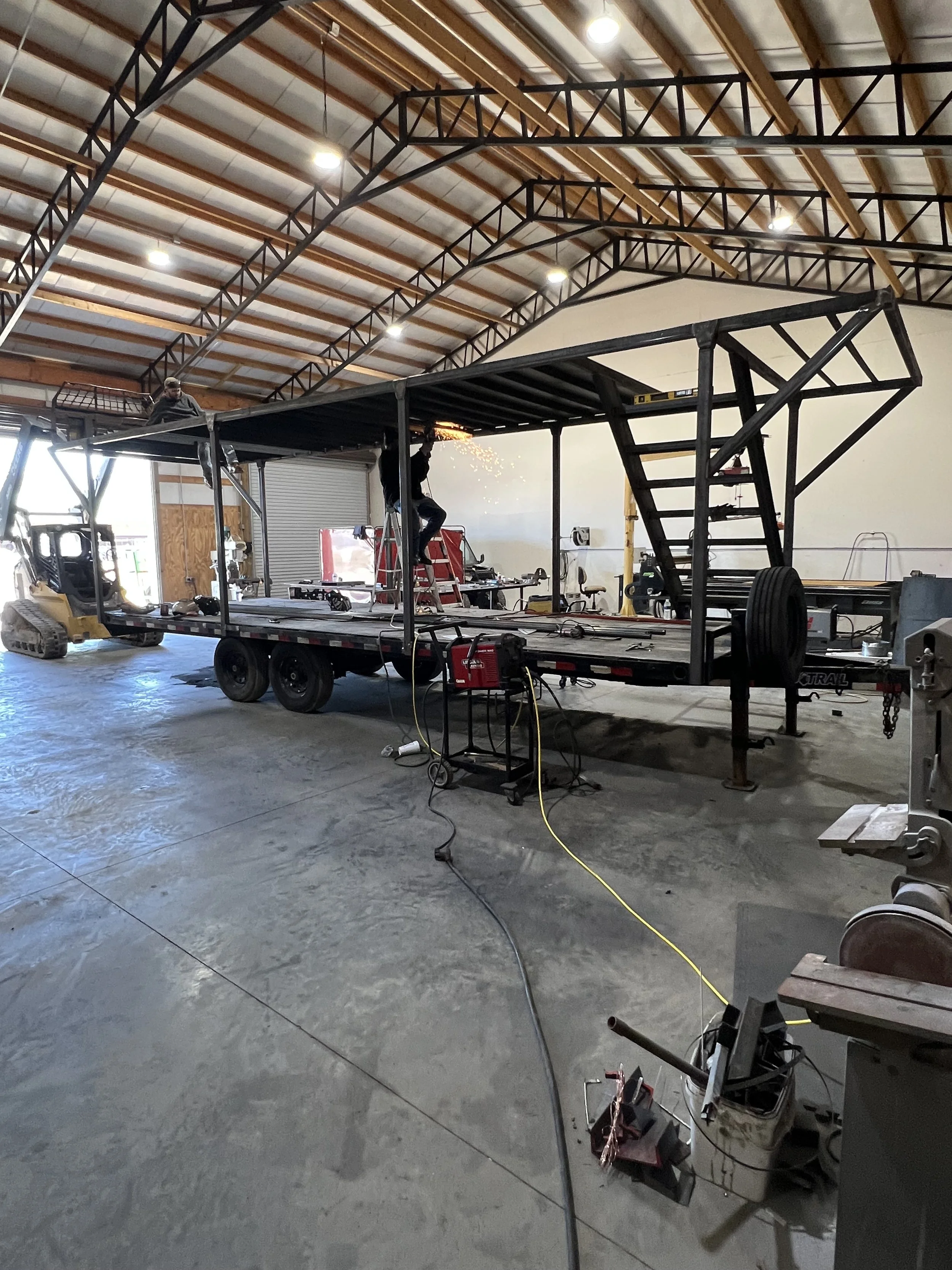Inside a warehouse or workshop with a large metal structure on a trailer, two workers welding with sparks flying, various equipment and tools scattered around.