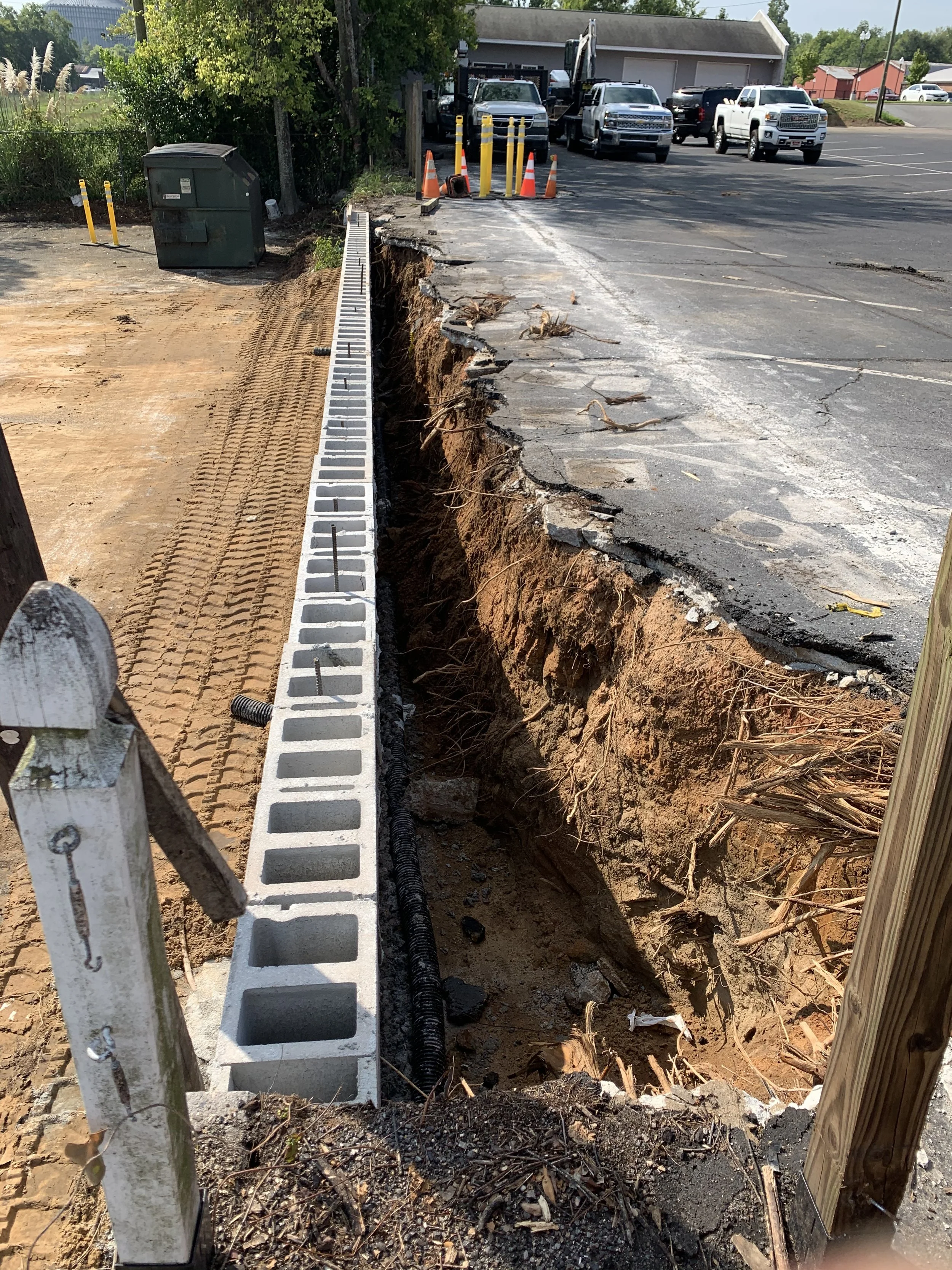 Construction site with a damaged section of a parking lot pavement and a trench next to a sidewalk. Orange traffic cones and barrier tape are set up nearby, with parked trucks and a building in the background.