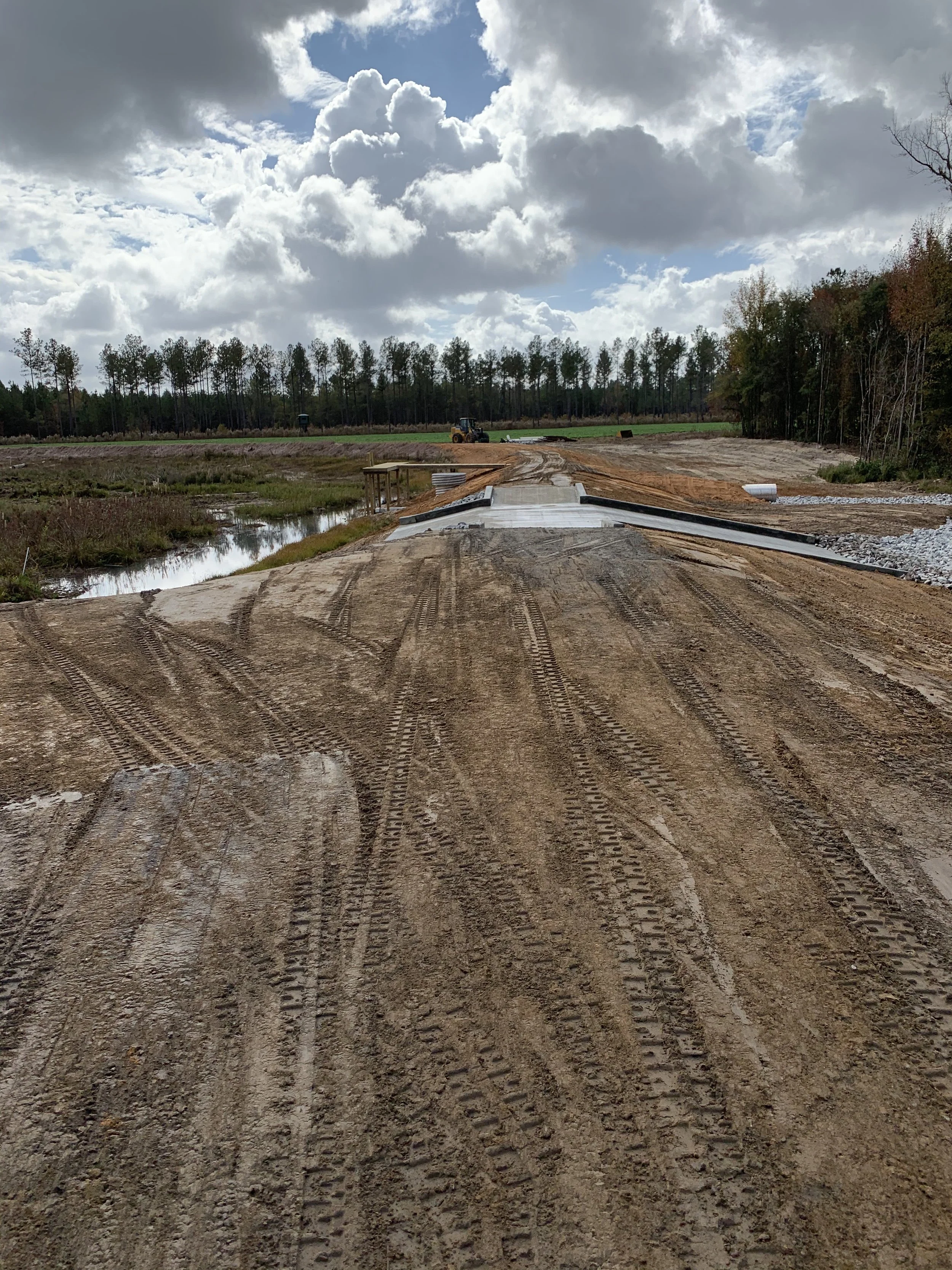 Construction site with a partially built road, tire tracks on dirt, machinery in the background, and cloudy sky.