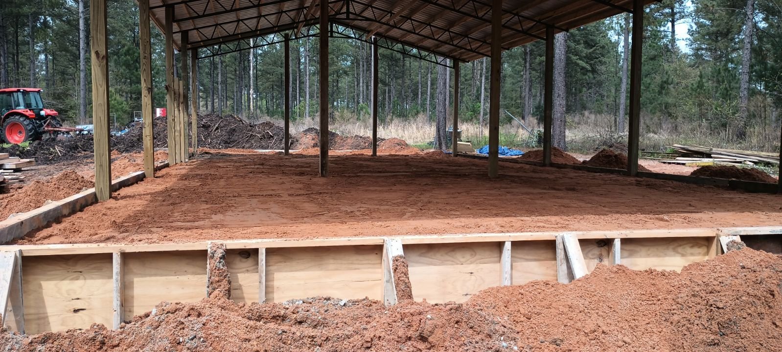 Construction site showing a cleared area prepared with a wooden frame for a building foundation, surrounded by trees and dirt mounds, with a red tractor on the left in a forested area.