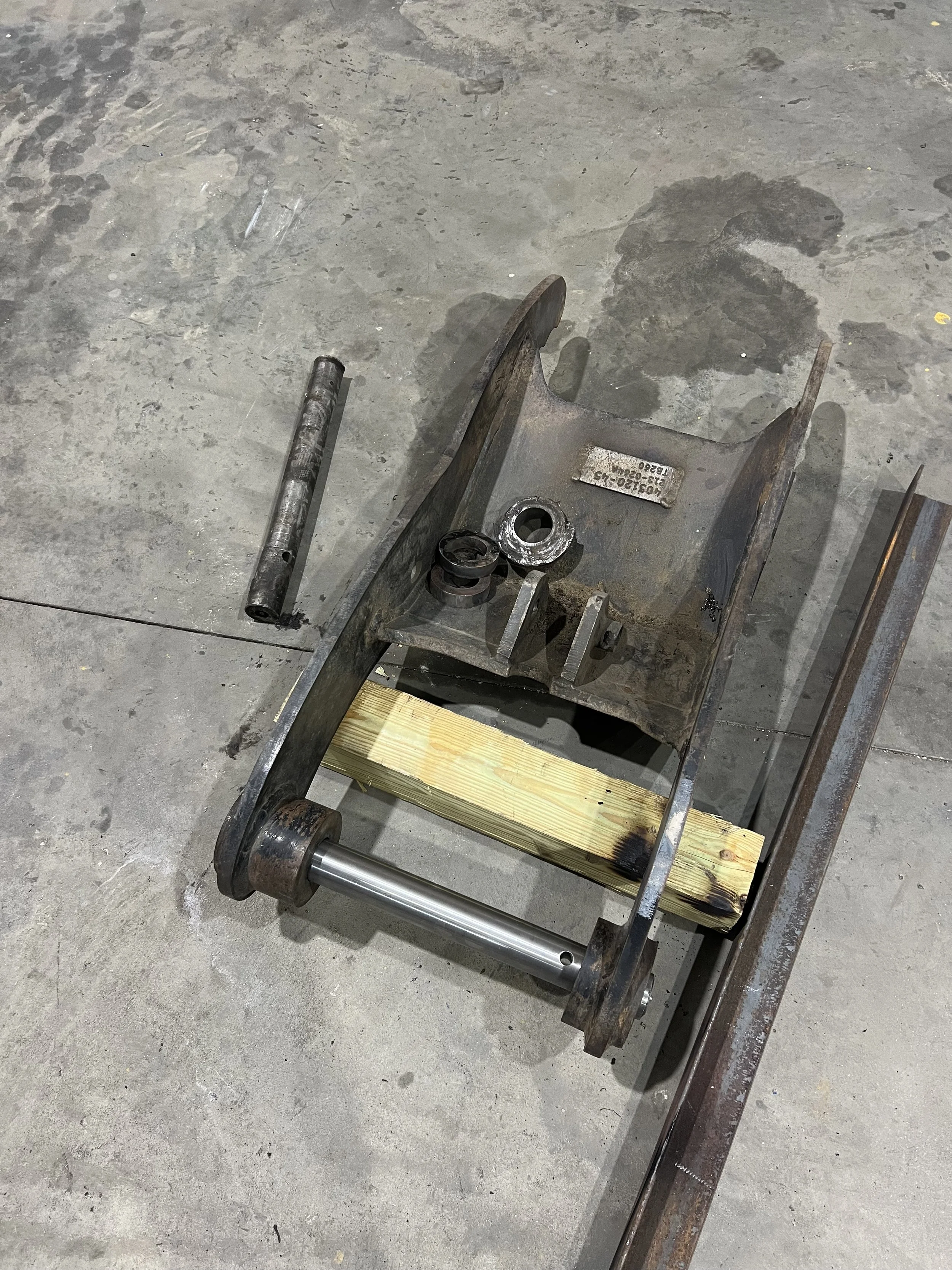 Disassembled metal machinery with a wooden block, metal rod, and various metal parts on a concrete floor.