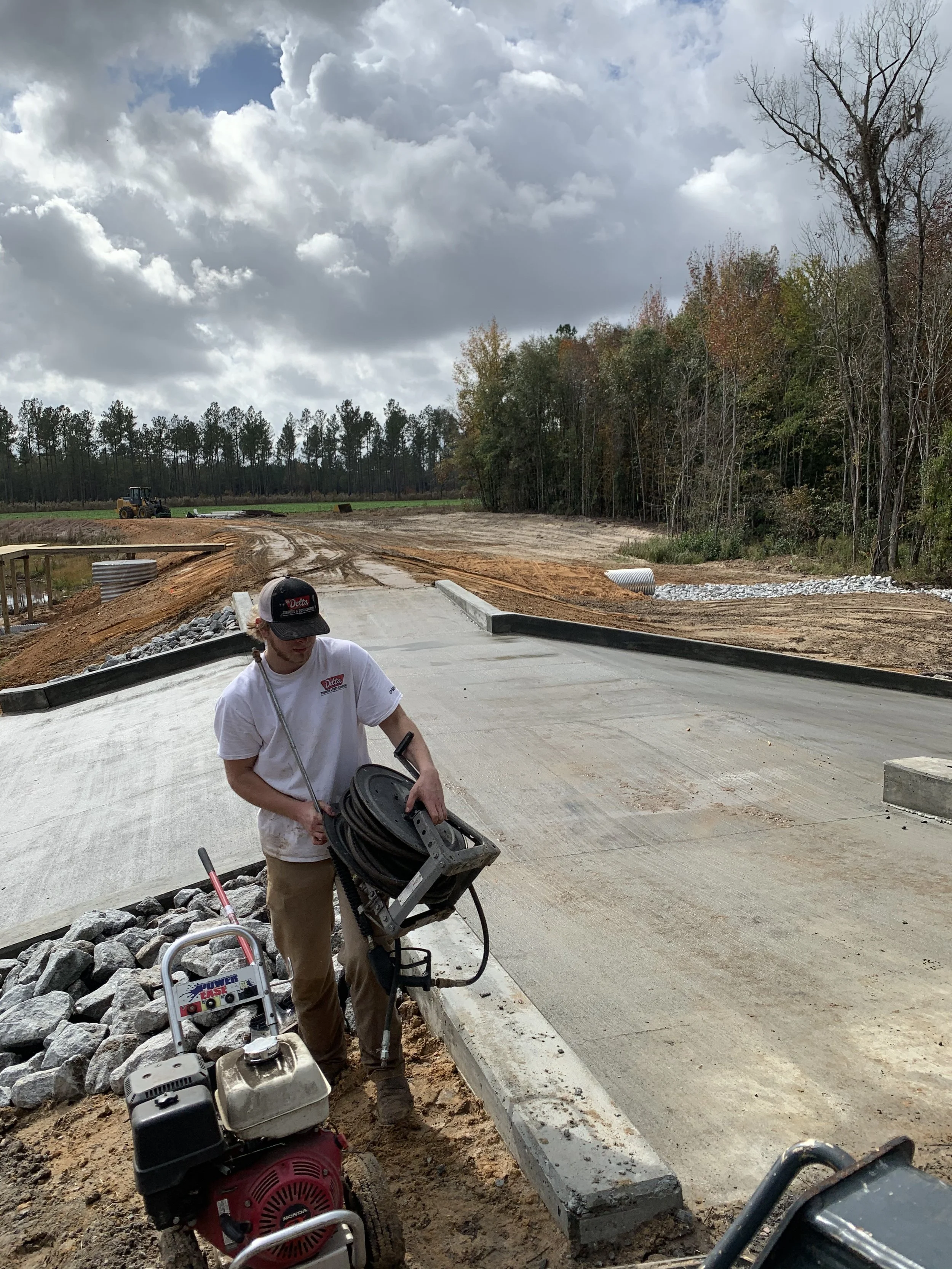 Construction worker preparing a concrete roadway with construction equipment on site, with cloudy sky and trees in the background.