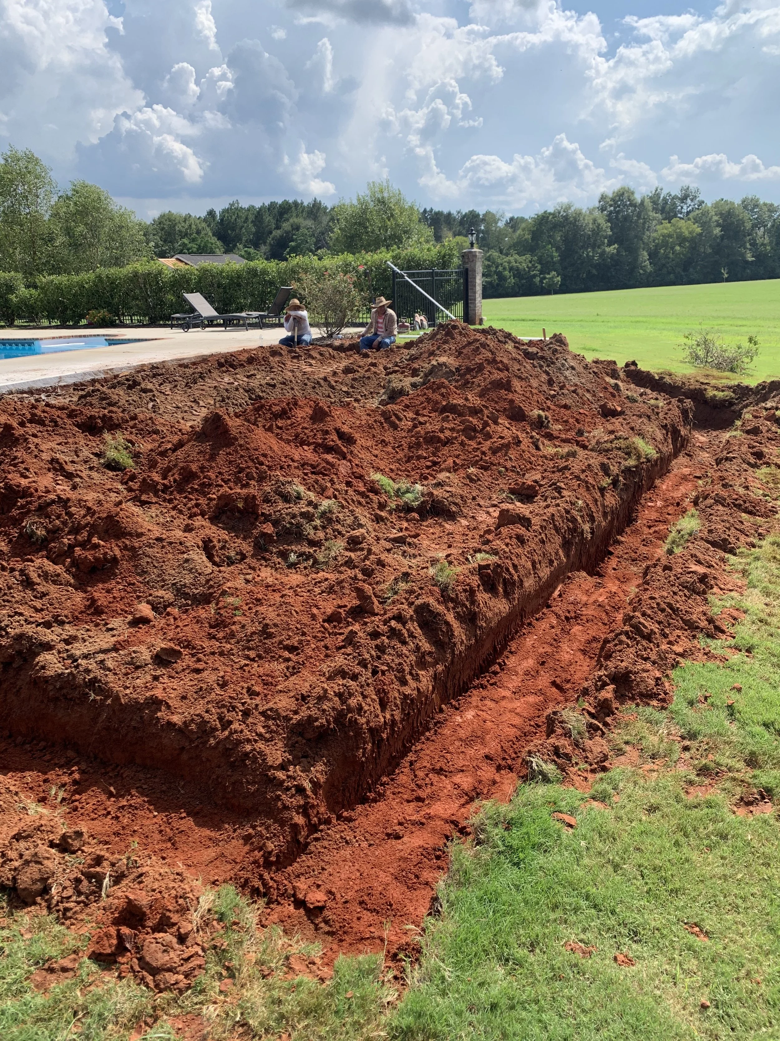 A freshly dug red dirt trench in a backyard with grass and trees, with two people sitting near the edge, and a pool with lounge chairs in the background under a partly cloudy sky.