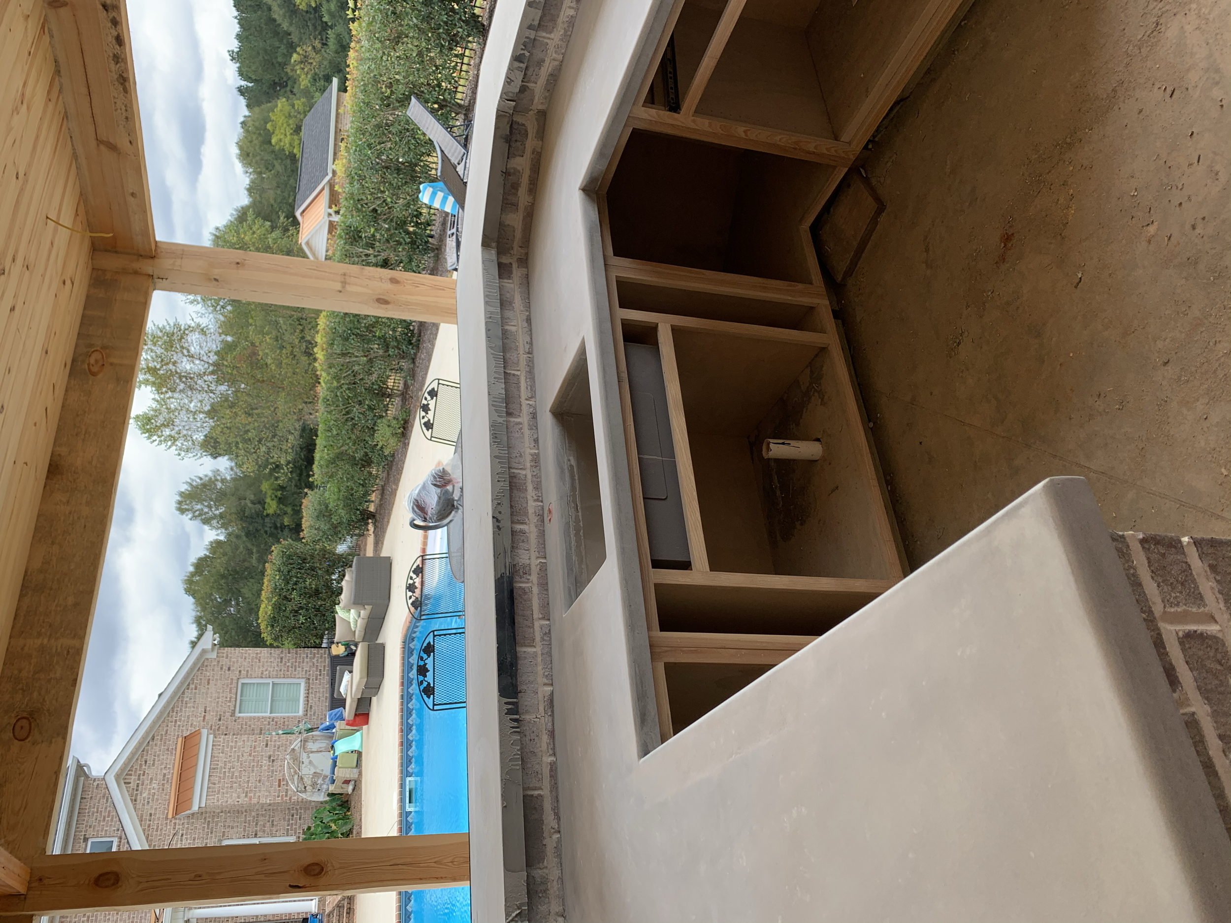 View from inside a house or shed under construction, showing unfinished wooden cabinets and an open wall leading to an outdoor backyard with a swimming pool, patio furniture, and surrounding trees.