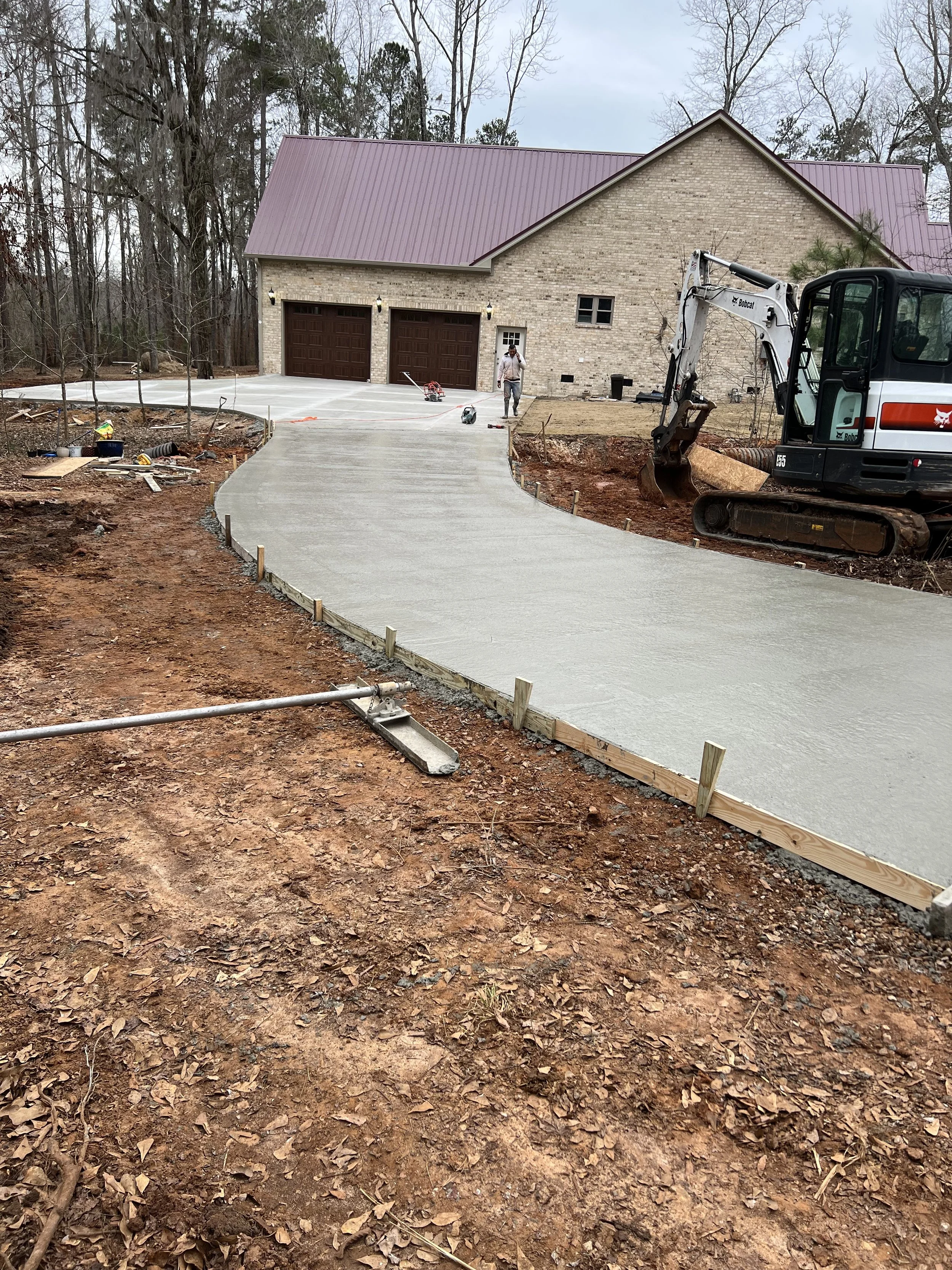 Construction site showing a newly poured concrete driveway leading to a house with a brick exterior and purple metal roof. There is construction equipment, including a small excavator, and workers working in the background.
