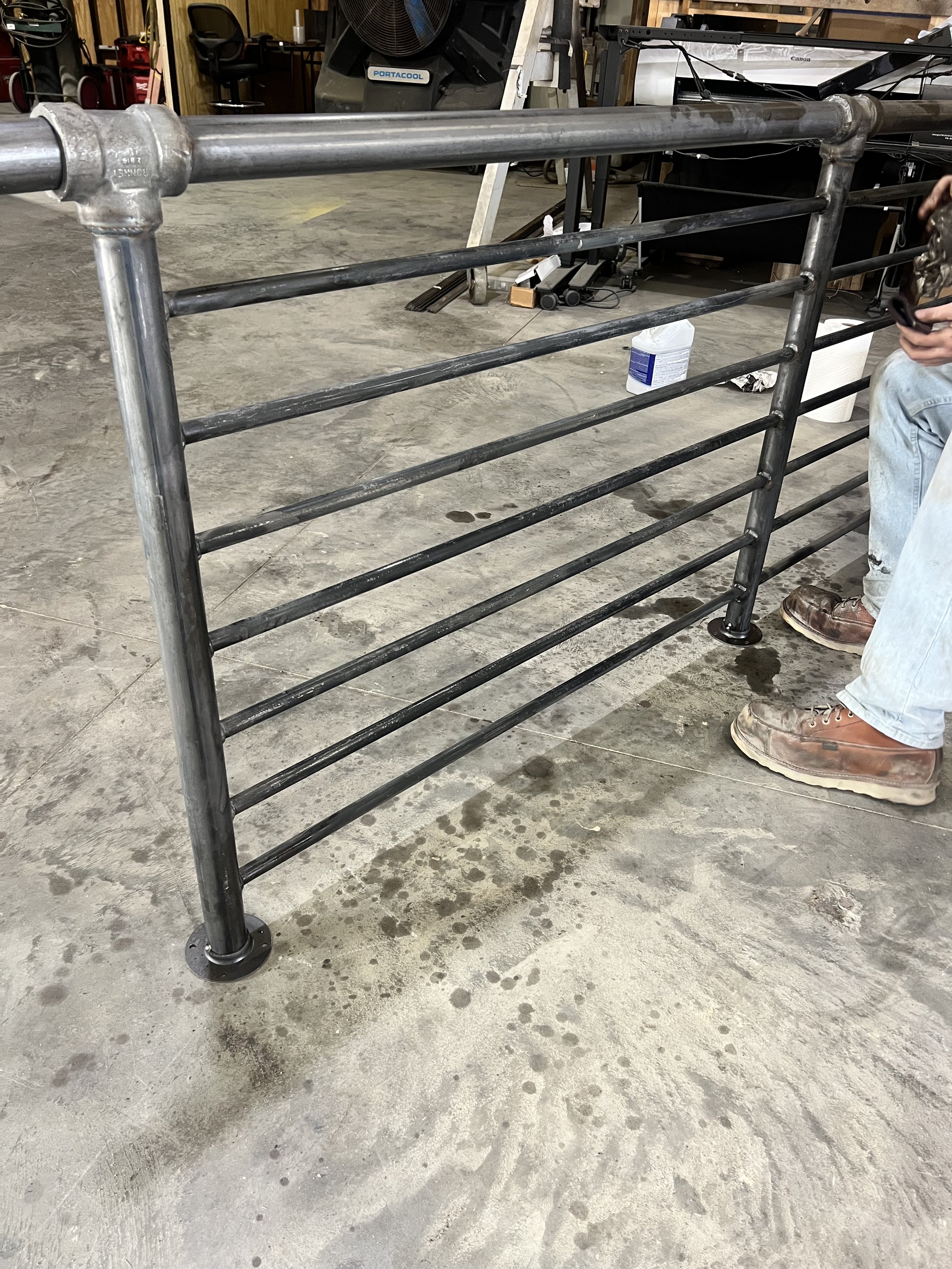Metal scaffold with horizontal bars in a workshop with concrete floor, tools, and a person wearing work boots sitting nearby.