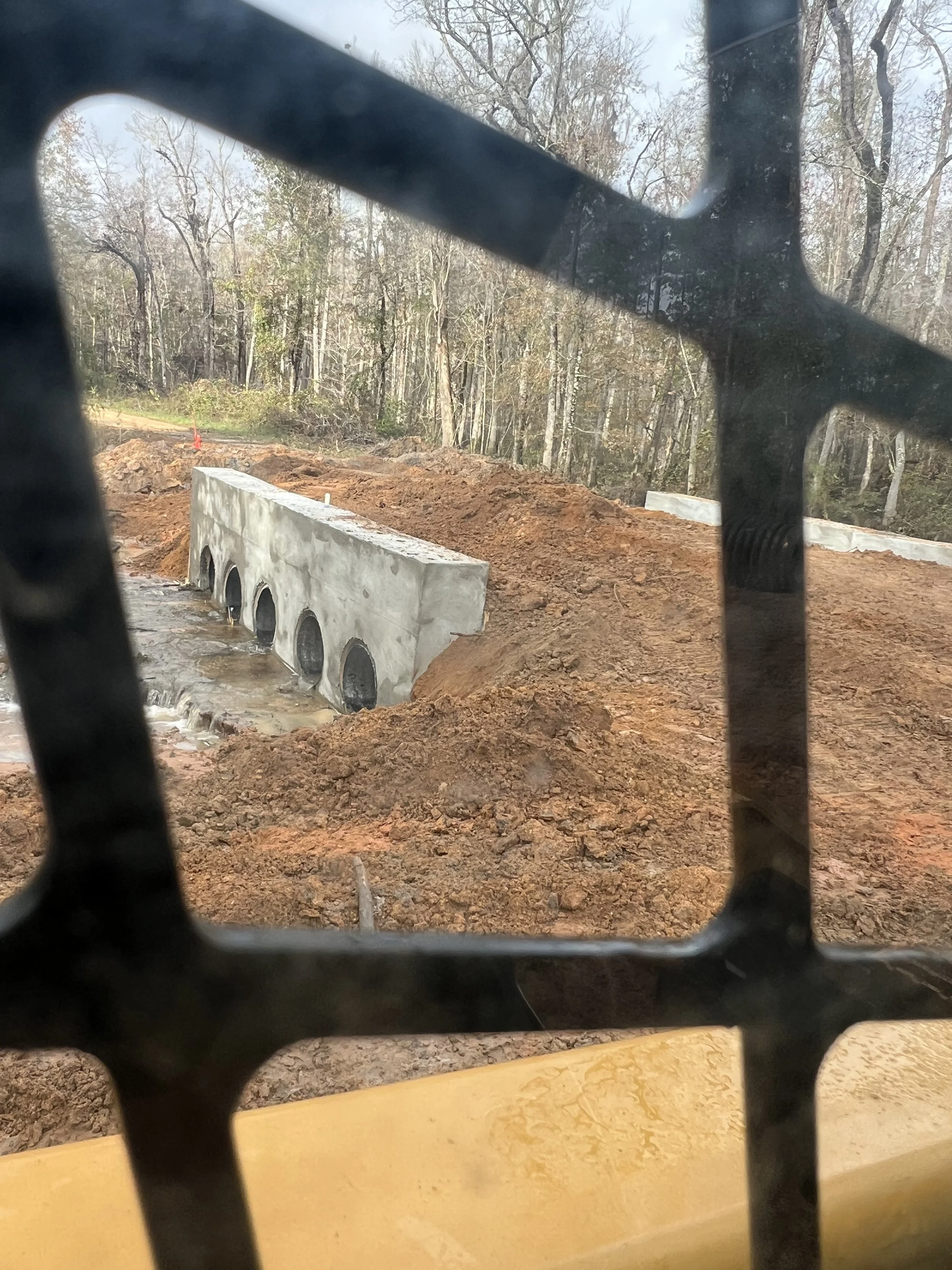 Construction site with a partially built concrete structure, surrounded by piles of dirt, seen through black metal fencing, with trees in the background.